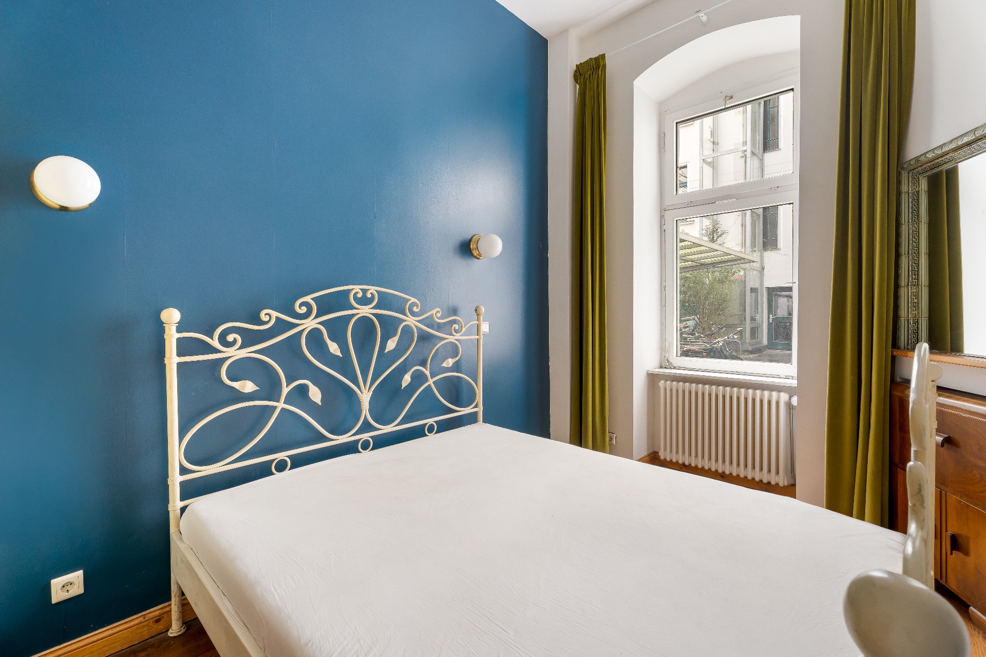 Schlafzimmer mit blau gestrichener Wand, weißem Metallbett und großem Fenster mit grünen Vorhängen.