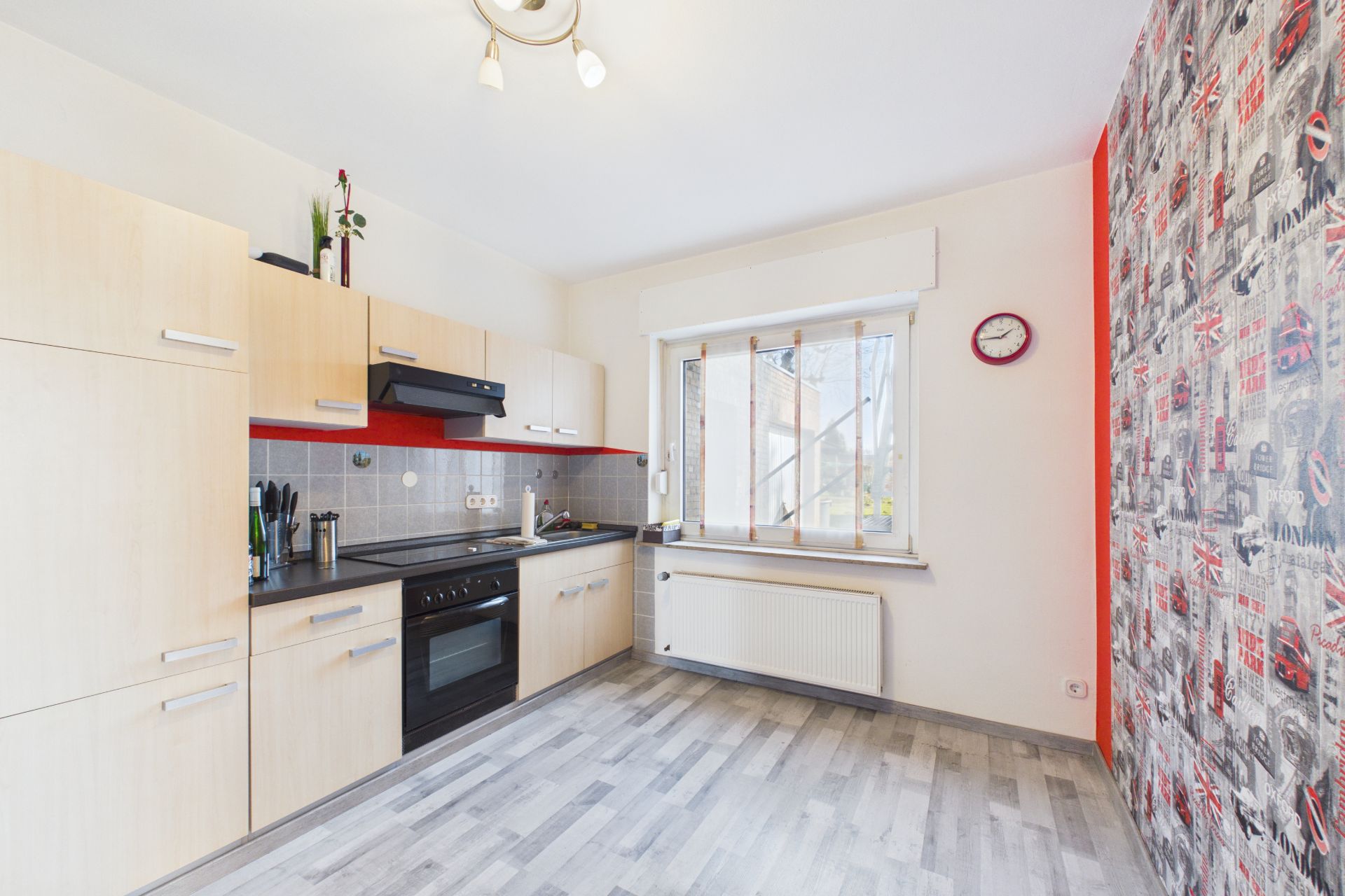 Helle Küche mit Holzfronten, großem Fenster, Uhr an der Wand und gemusterten Tapeten.