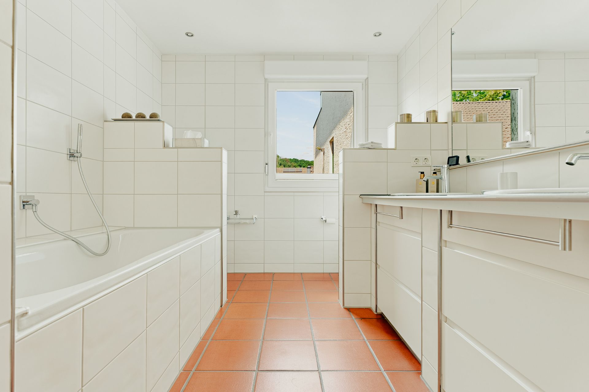 Modernes Badezimmer mit Badewanne, weißen Fliesen und großem Spiegel, terrakottafarbenem Boden.