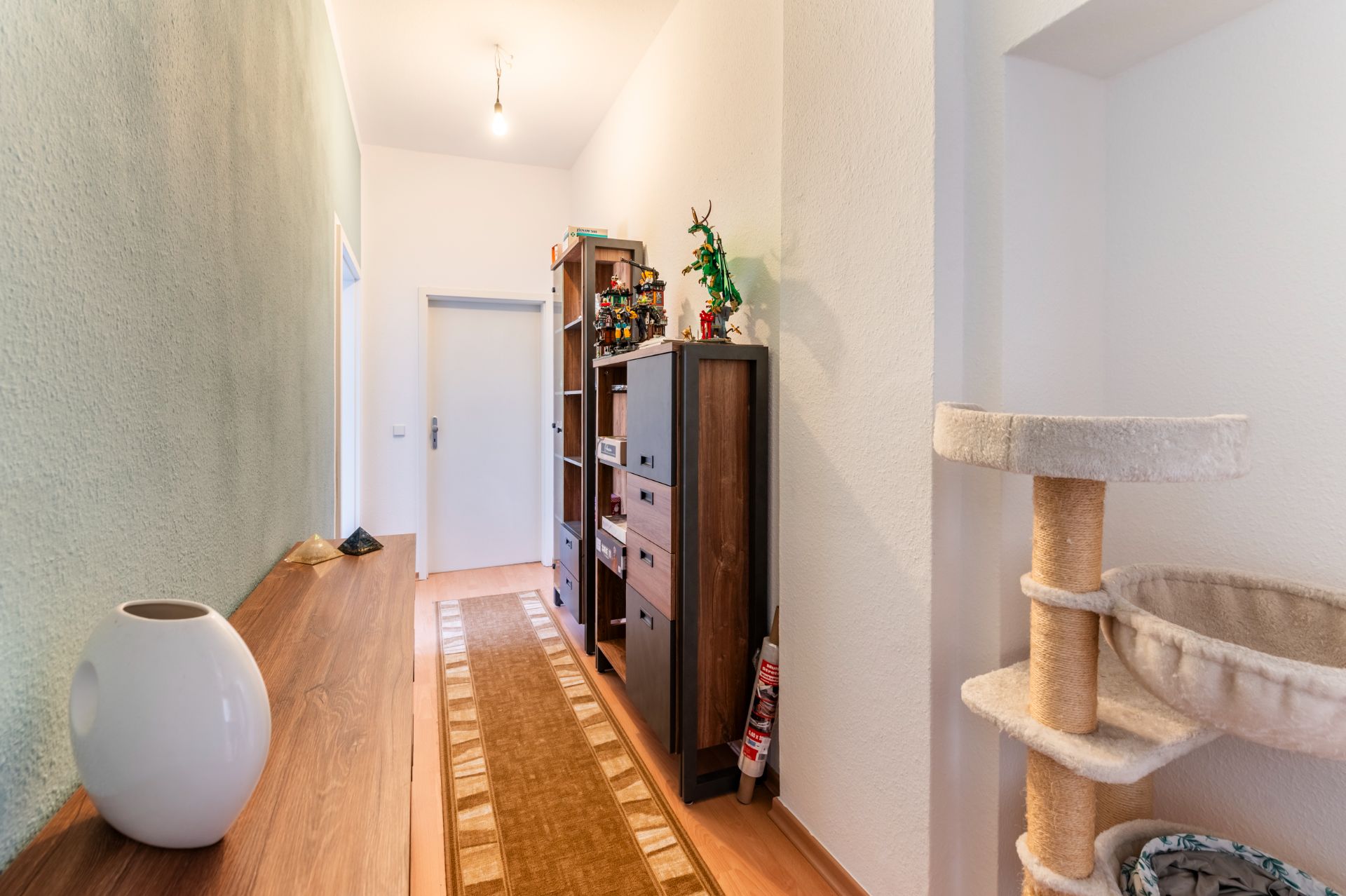 Heller Flur mit Teppich, Regal, Kommode, Kratzbaum und dekorativer Vase auf einem Holztisch.
