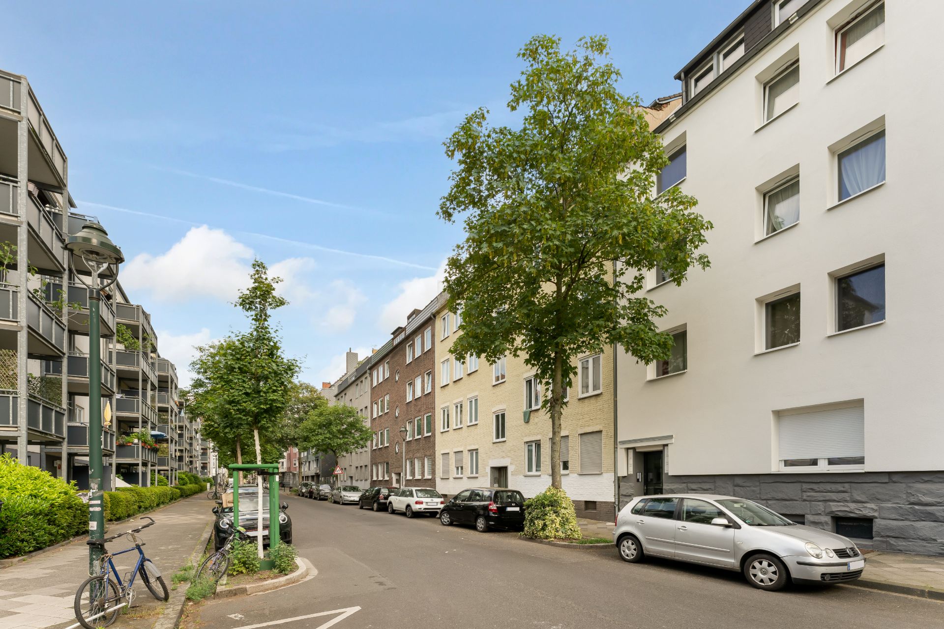 Wohnstraße mit parkenden Autos, Fahrrädern und mehrstöckigen Wohngebäuden bei Tageslicht.