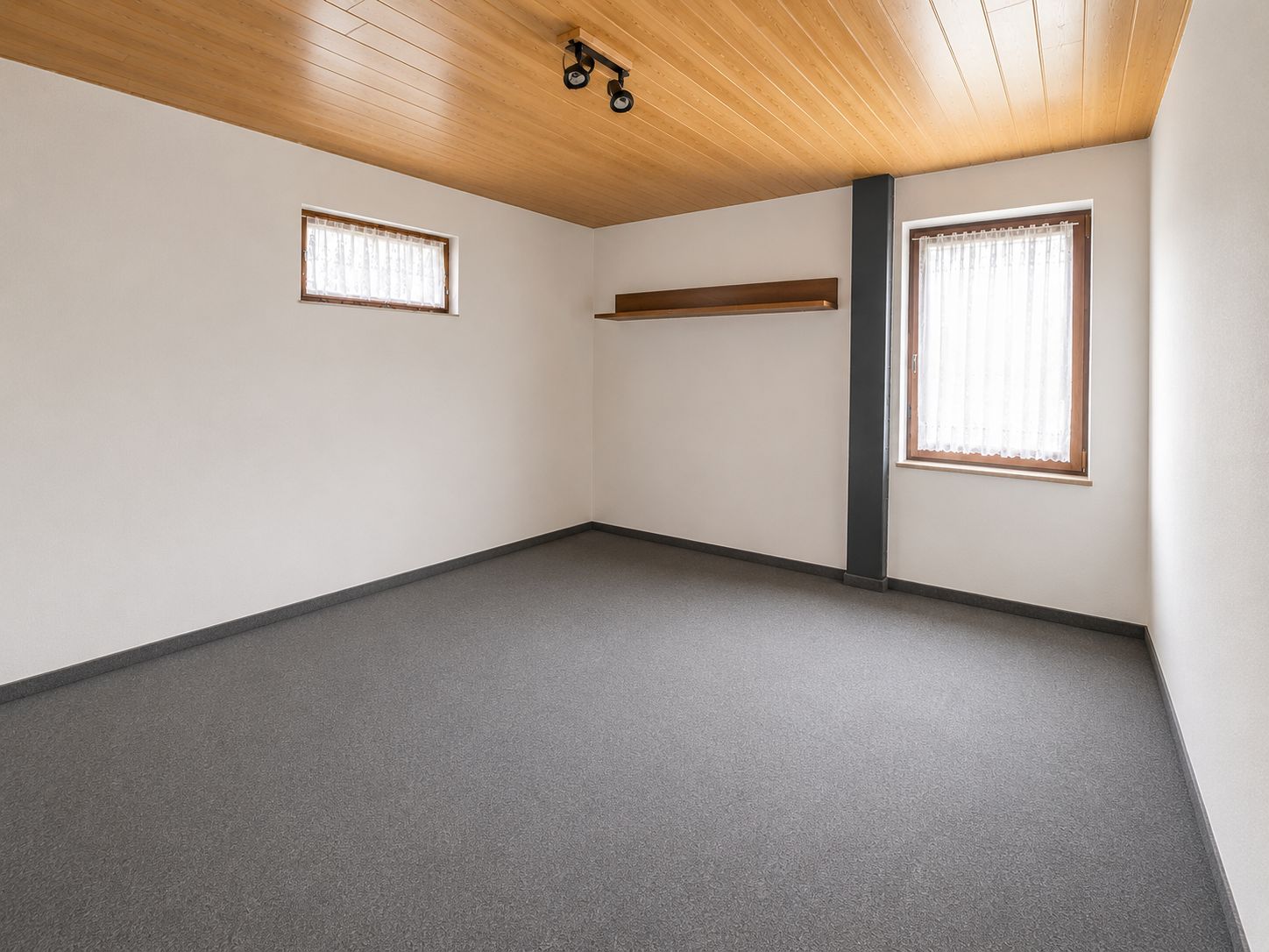 Leeres Zimmer mit Teppichboden, Holzdecke, zwei Fenstern und einem Wandregal.