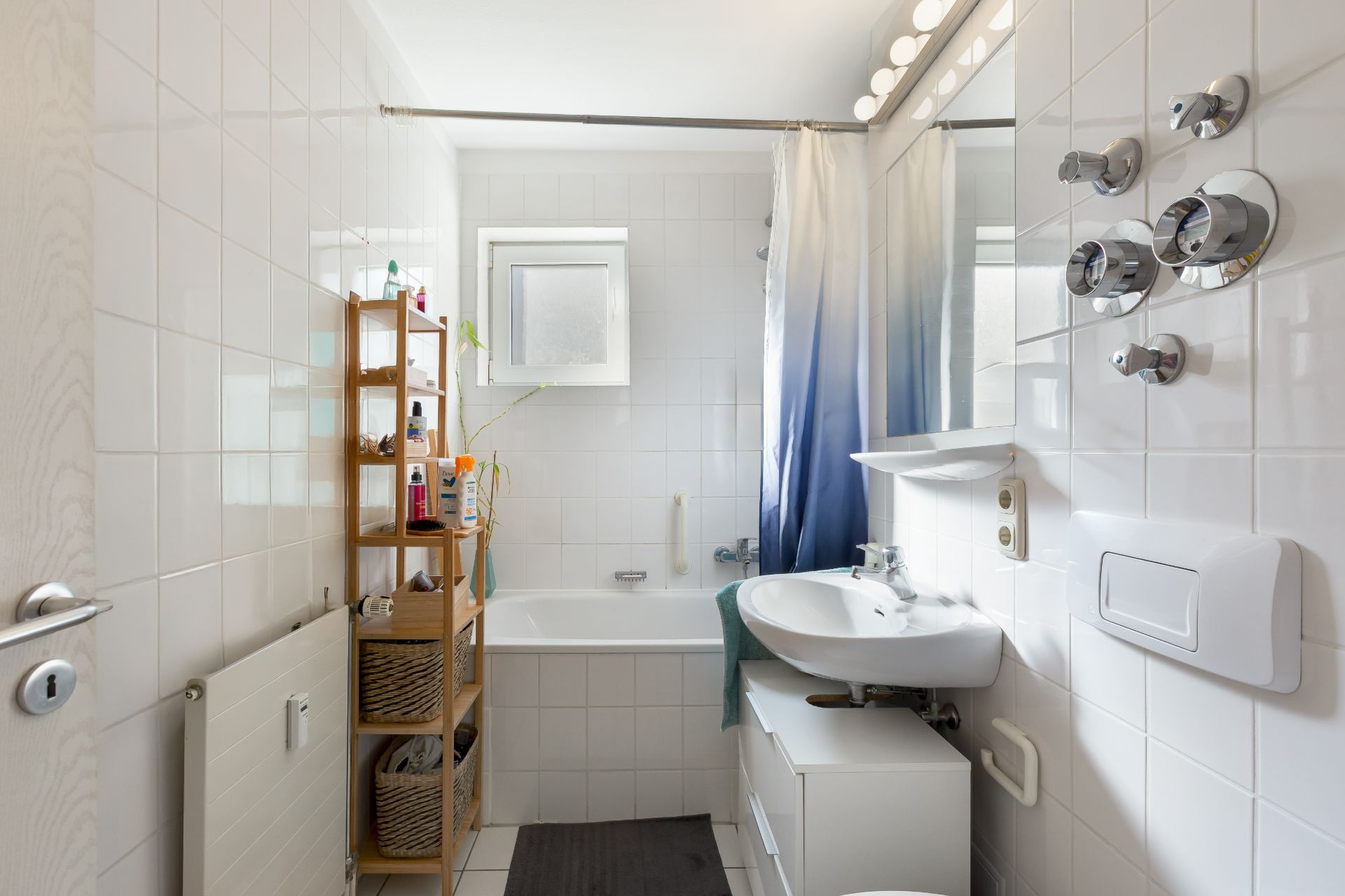 Helles Badezimmer mit Badewanne, Waschbecken, Regal und weißen Fliesen.