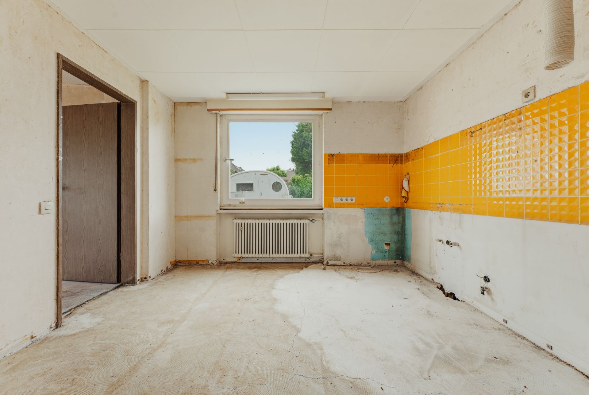 Leerer Raum mit gelben Wandfliesen, Fenster und Heizkörper, sichtbare Anschlüsse an der Wand.