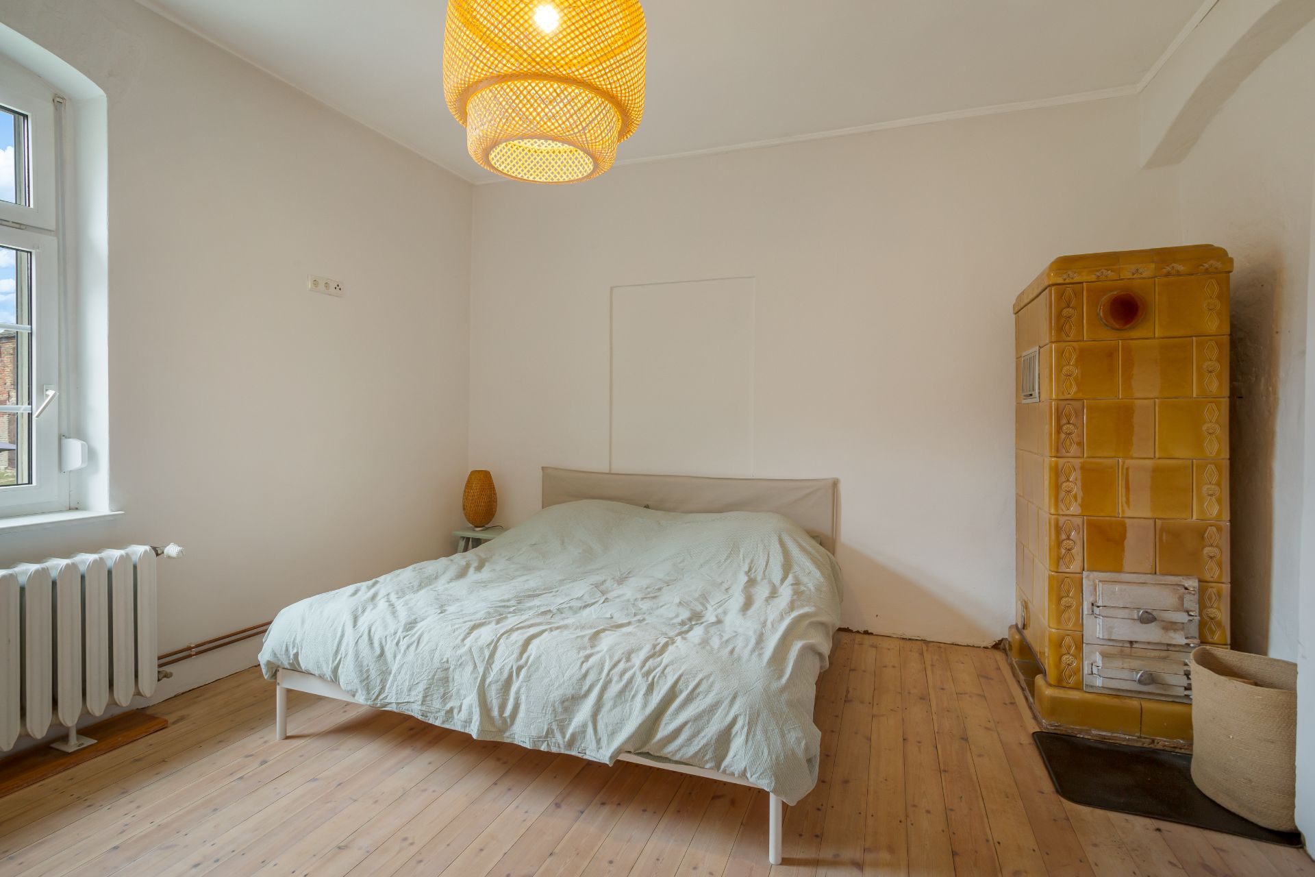 Schlafzimmer mit Doppelbett, Nachttisch, Fenster, Heizkörper und gelbem Kachelofen.
