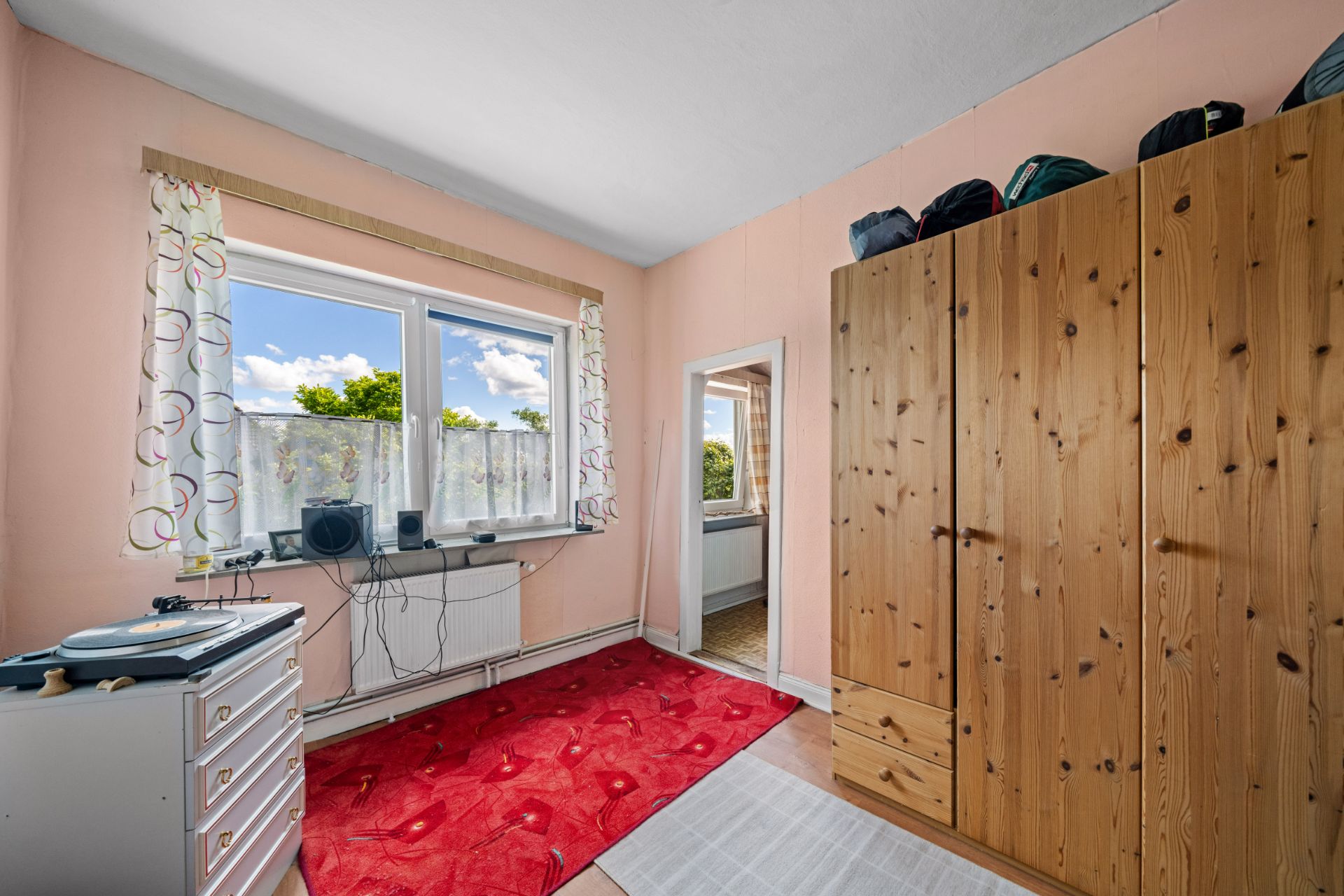 Kleines Zimmer mit Holzgarderobe, rotem Teppich, Kommode und Fenster mit Blick nach draußen.
