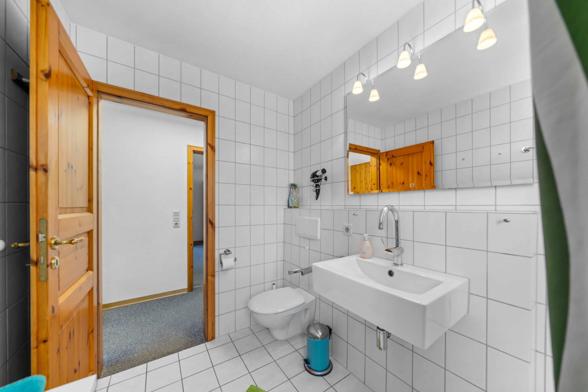 Helles Bad mit weiß gefliesten Wänden, Wand-WC, Waschbecken und großem Spiegel.
