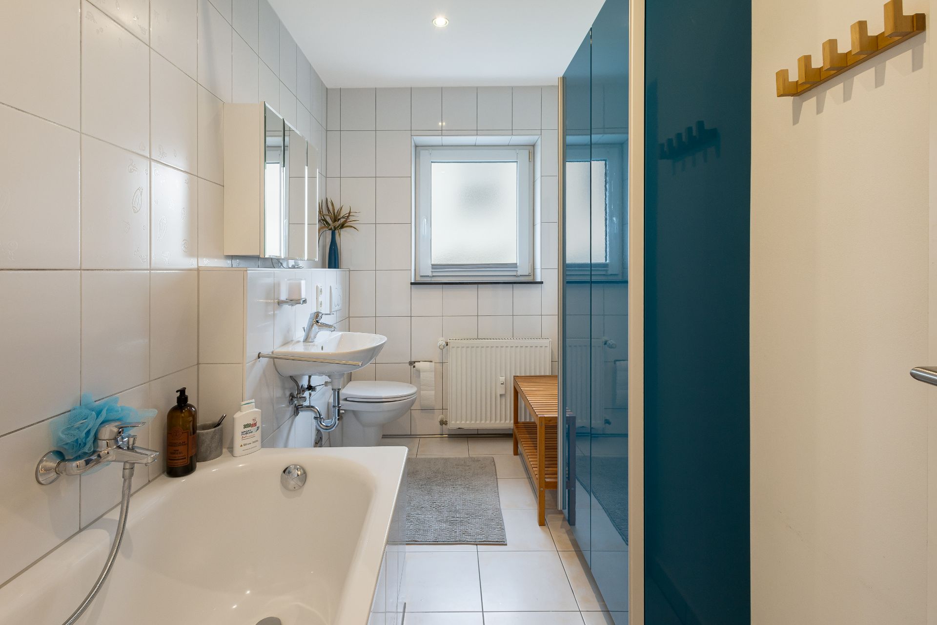 Helles Badezimmer mit Badewanne, Waschbecken, Toilette, Fenster und Holzbank.