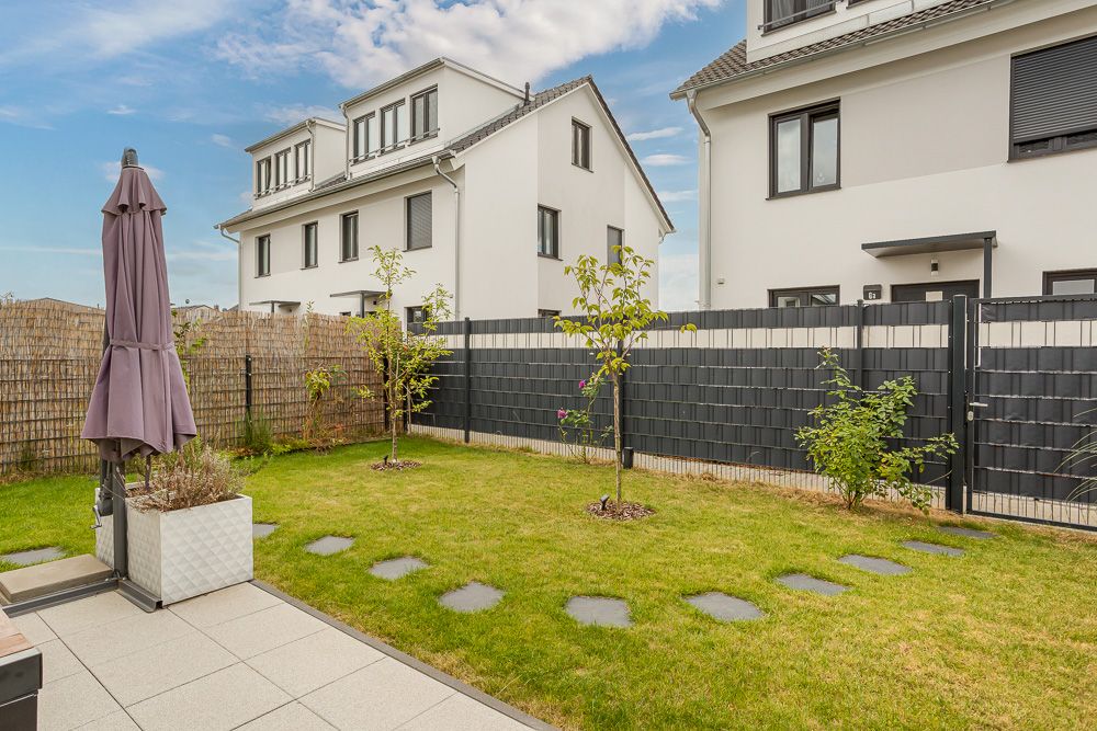 Gepflegter Garten mit Rasen, kleinen Bäumen, Terrasse und Sichtschutzzaun vor Wohnhäusern.