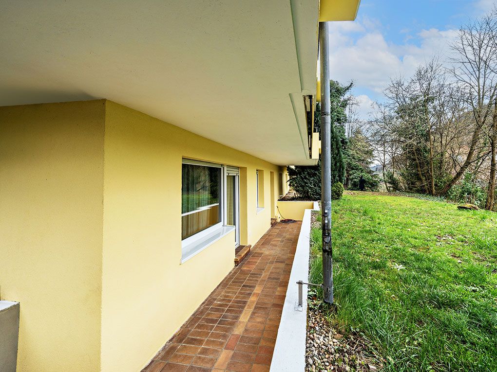 Überdachte Terrasse mit gelber Fassade, braunen Fliesen und angrenzendem Garten.