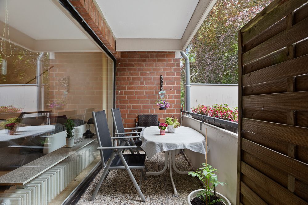 Überdachter Balkon mit Tisch, Stühlen, Blumen und Pflanzen, angrenzend an Wohnzimmer mit Glasfront.