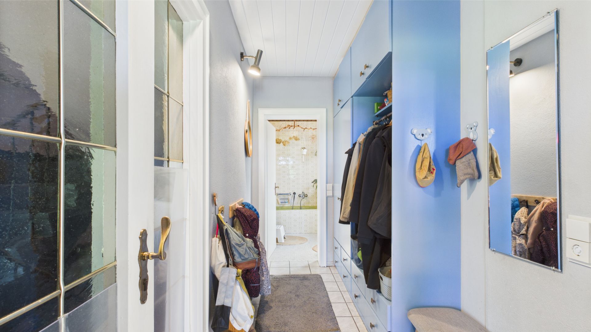 Heller Flur mit Garderobe, Spiegel, Kleiderhaken und Bank, Blick ins Badezimmer.