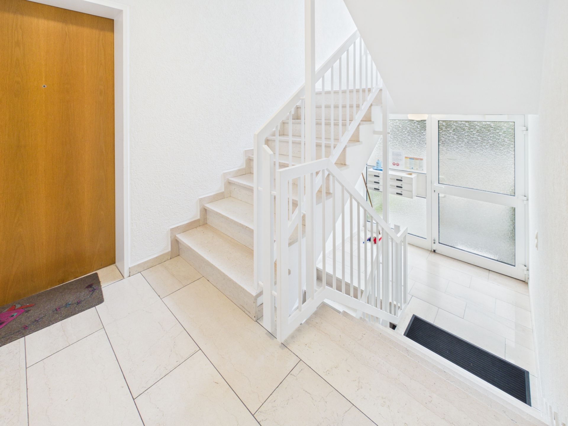 Heller Treppenhausbereich mit weißen Wänden, Fliesenboden und Holztür.