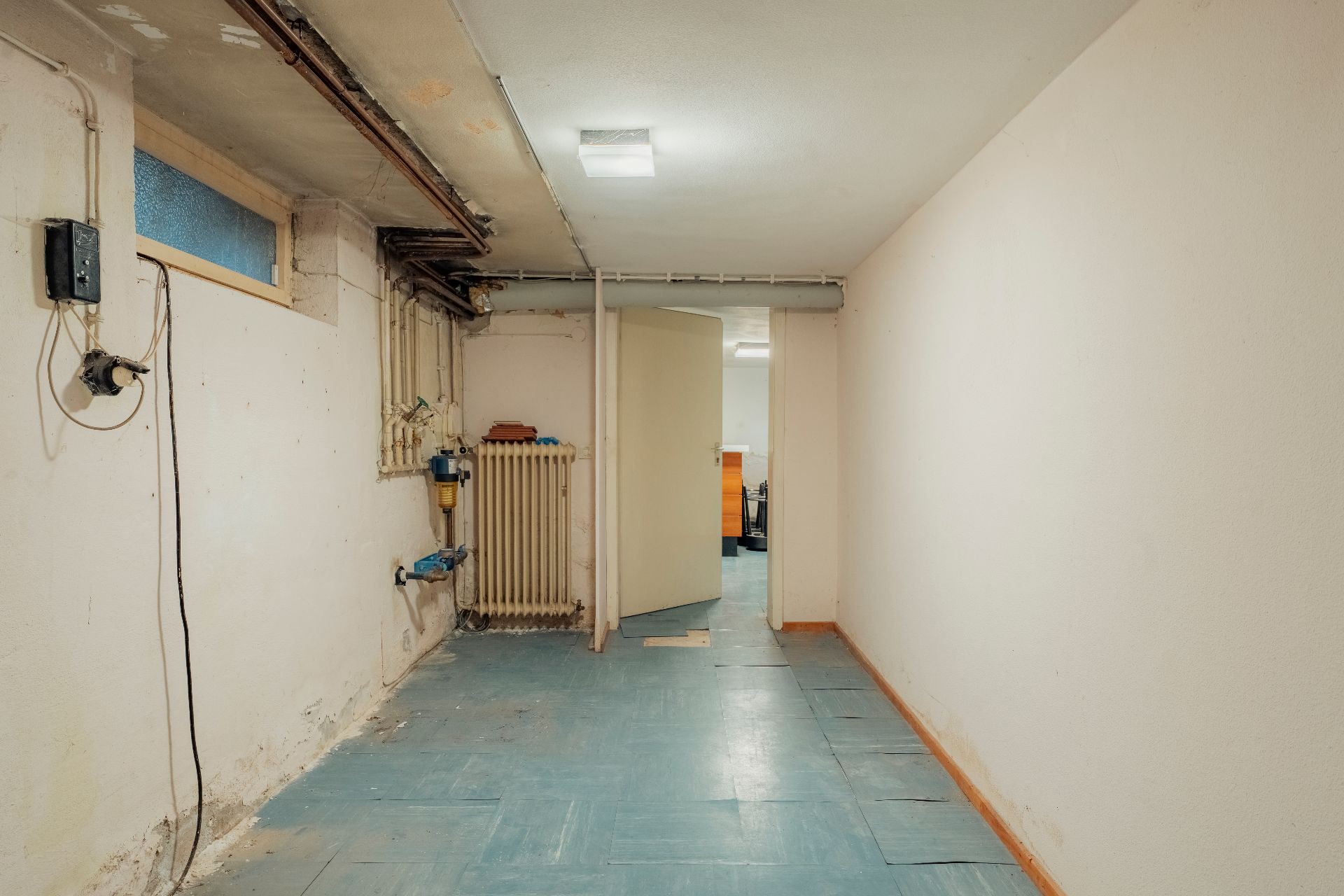 Langer, leerer Kellerraum mit sichtbaren Rohren und Heizung, Tür zum nächsten Raum offen.