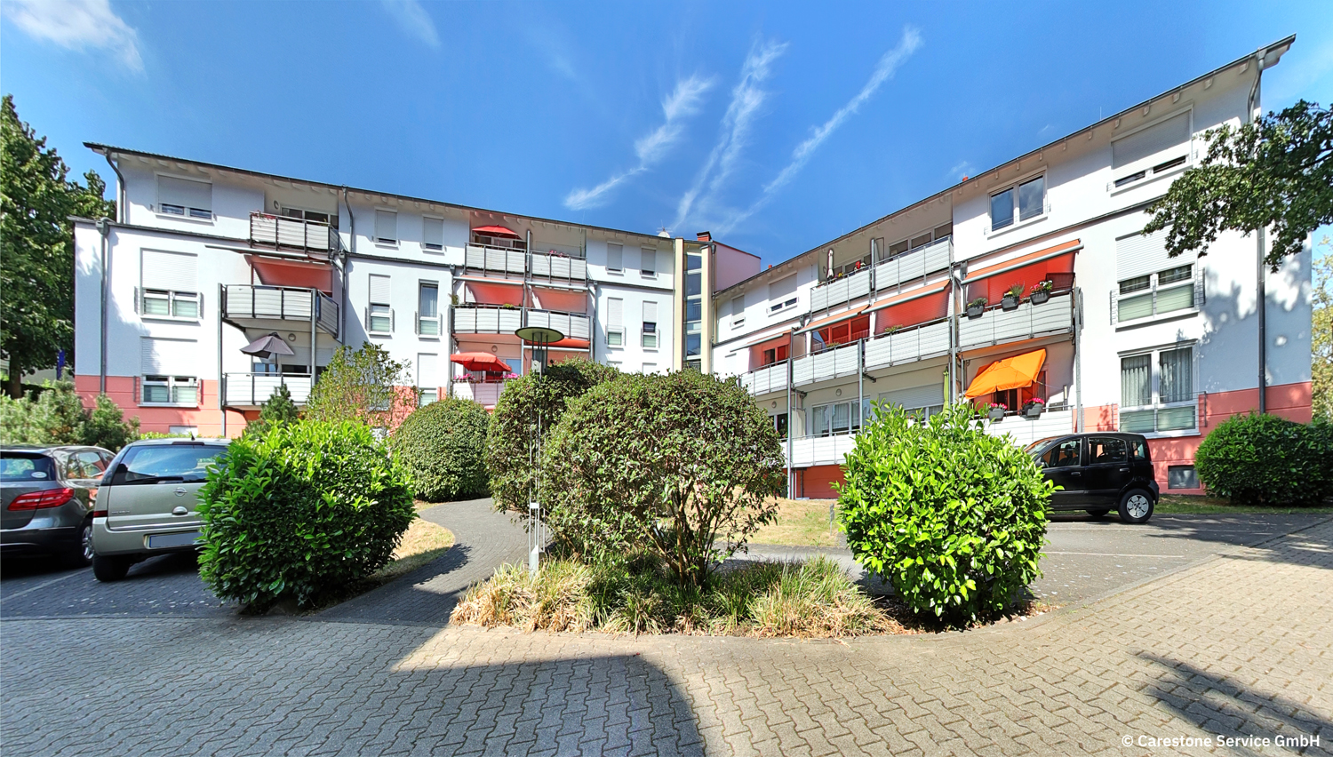 Mehrfamilienhaus mit Balkonen, geparkten Autos und Büschen im Innenhof bei sonnigem Wetter.