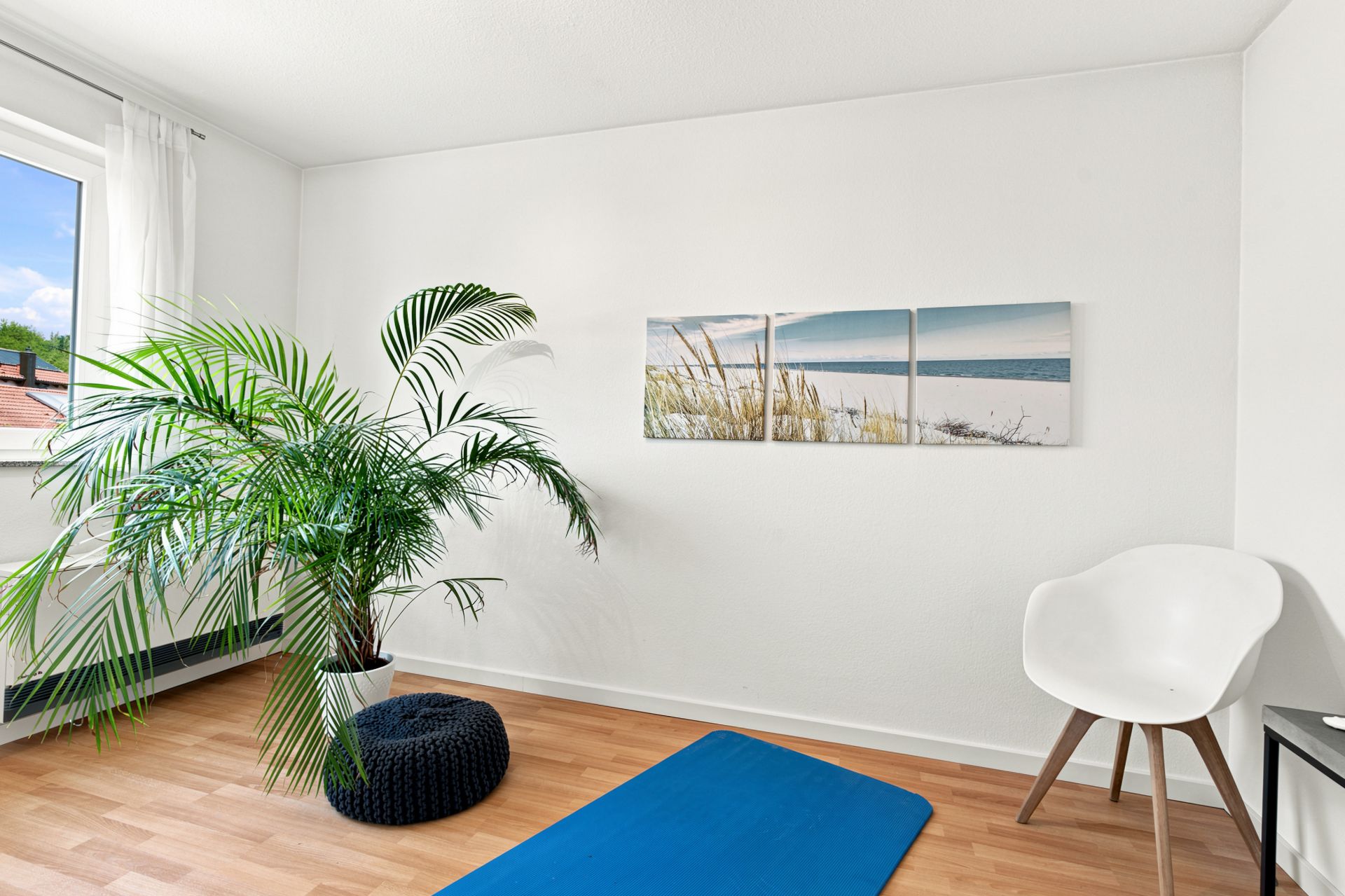 Helles Zimmer mit großem Fenster, Zimmerpflanze, weißem Stuhl und blauer Matte auf Holzboden.