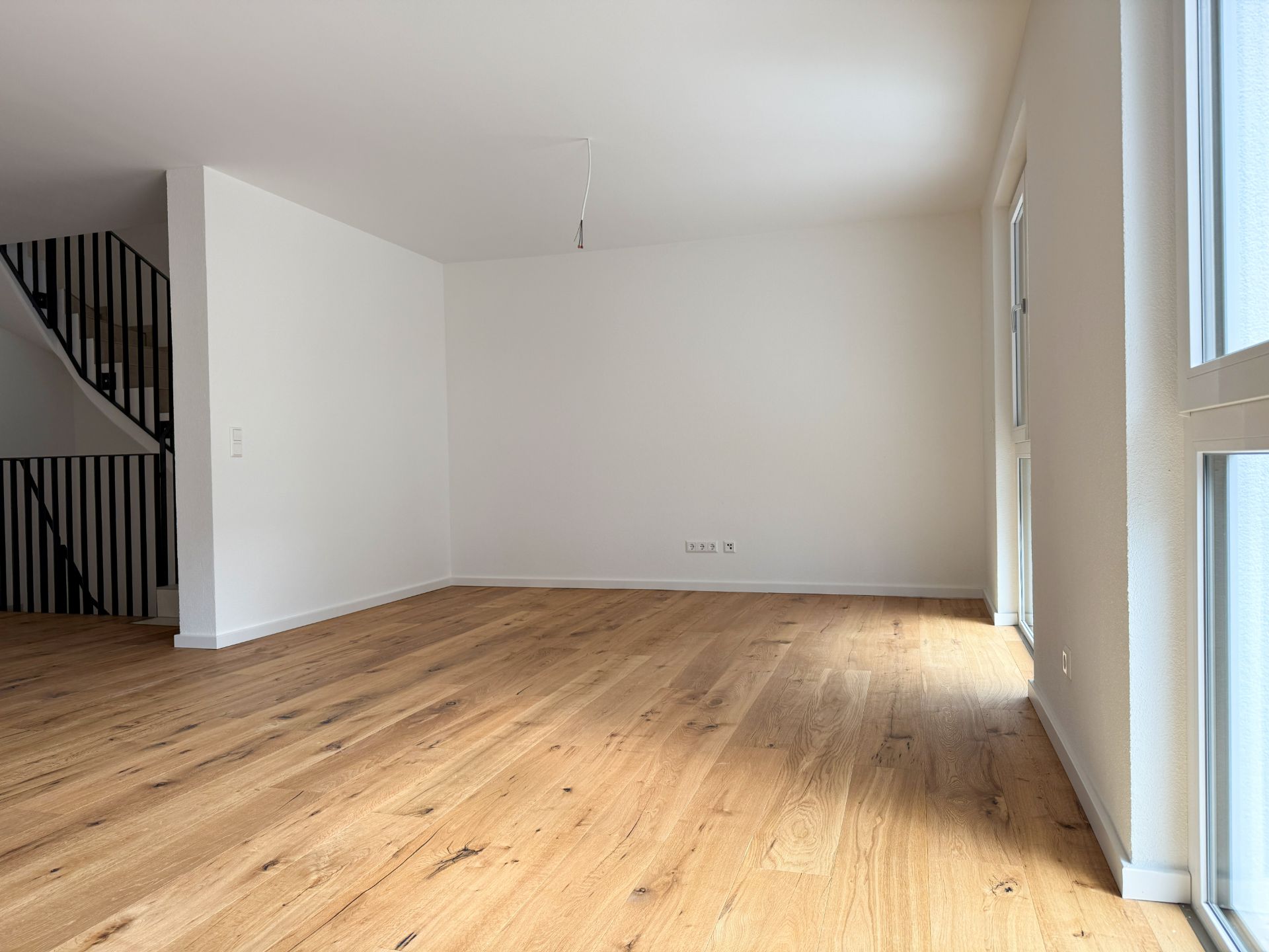 Helles, leeres Wohnzimmer mit Holzfußboden und großen Fenstern, links eine Treppe mit schwarzem Geländer.