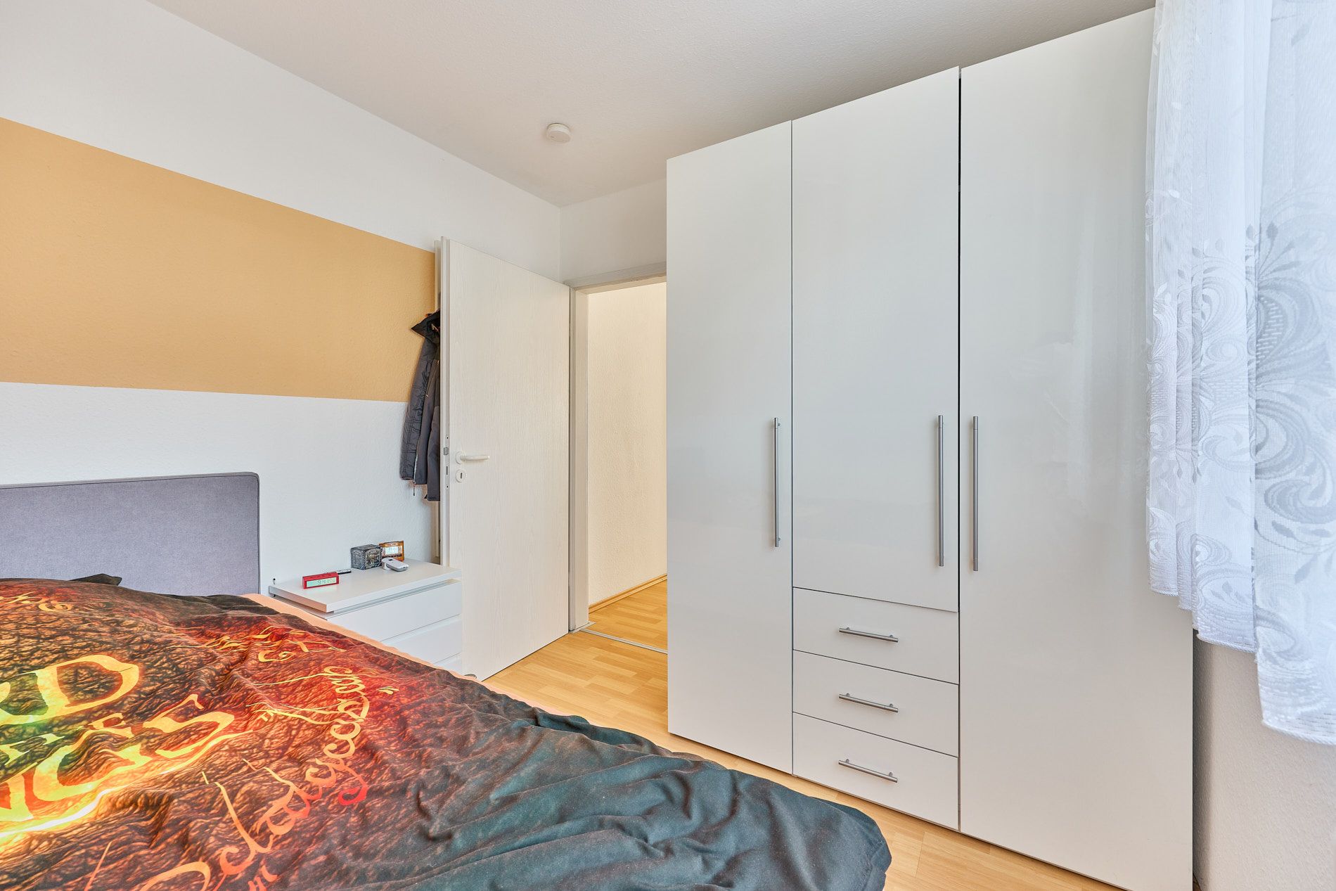 Modernes Schlafzimmer mit weißem Kleiderschrank, Bett und einem Fenster mit Gardine.