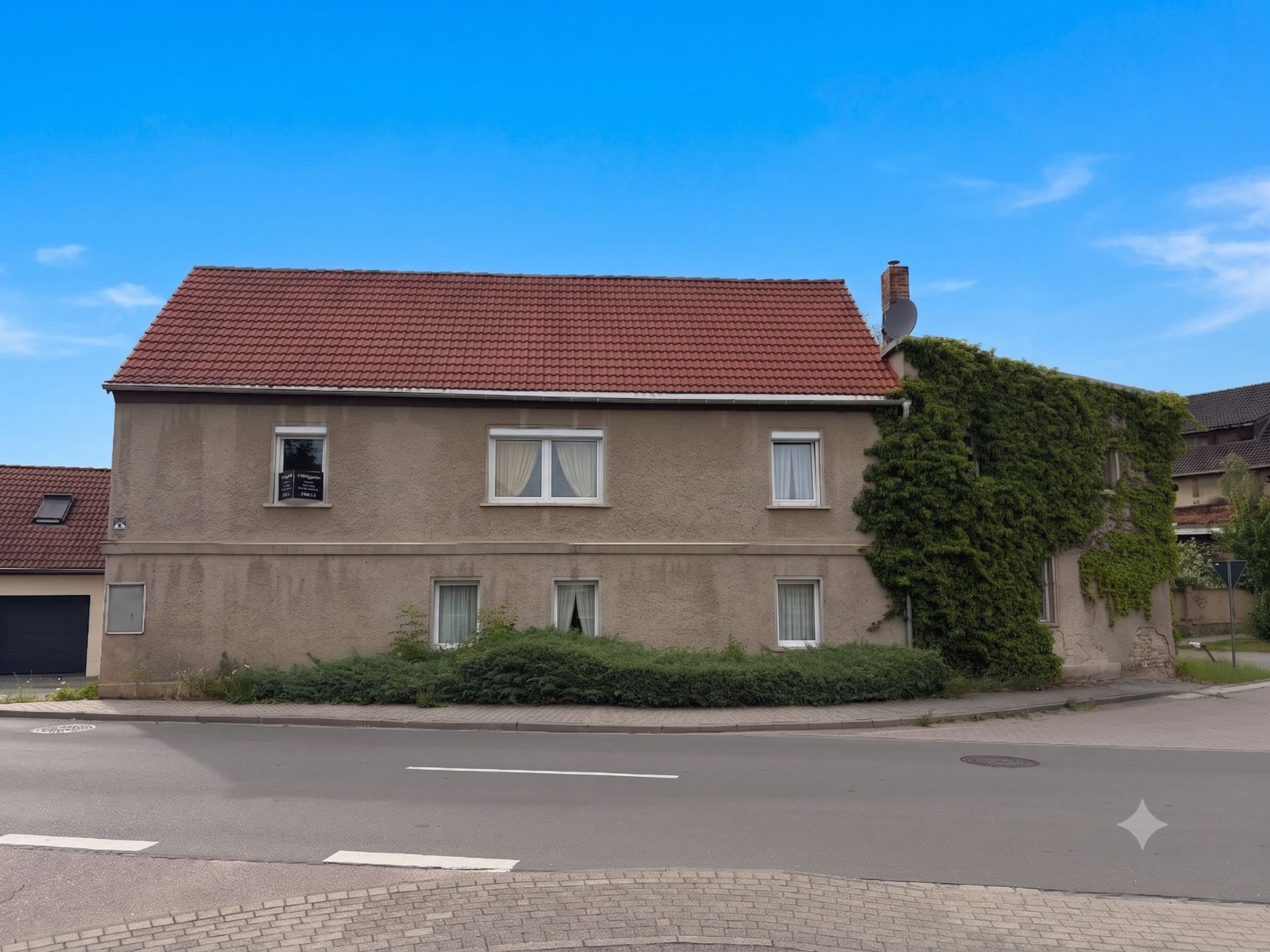 Zweistöckiges Haus mit rotem Dach, begrüntem Seitenbereich und mehreren Fenstern an einer Straße.
