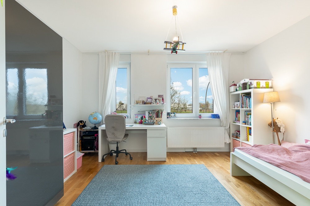 Helles Kinderzimmer mit Schreibtisch, Bett, Regal und zwei Fenstern.