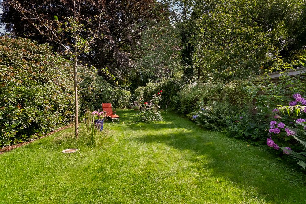 Langer Garten mit Rasenfläche, Bäumen, Sträuchern, Blumen und einer roten Gartenliege.