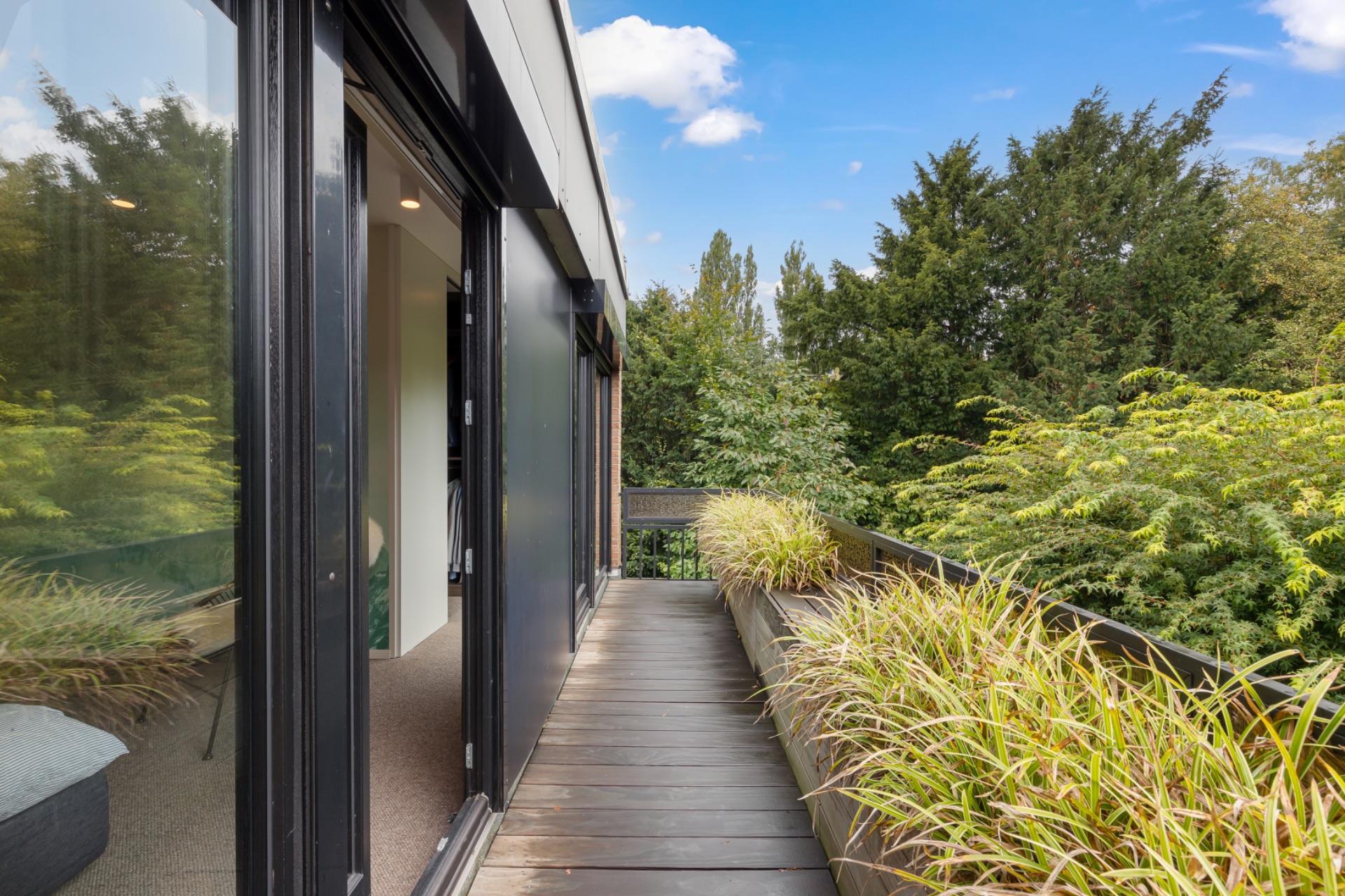 Balkon mit Holzboden, Pflanzenkübeln und Blick auf grüne Bäume, Zugang durch große Glasschiebetüren.