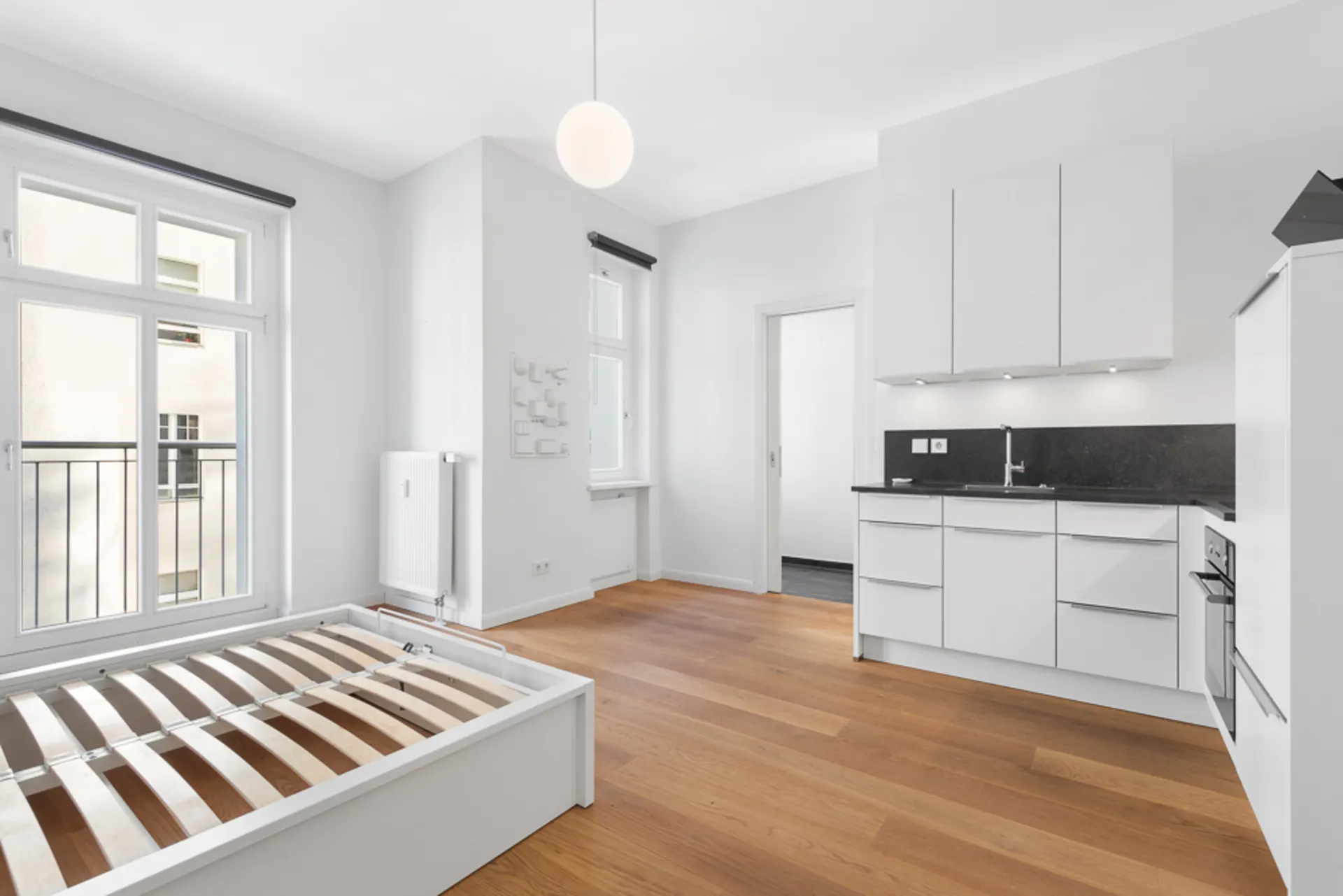 Helles Zimmer mit Holzboden, Küchenzeile und einem Bettgestell vor zwei Fenstern.
