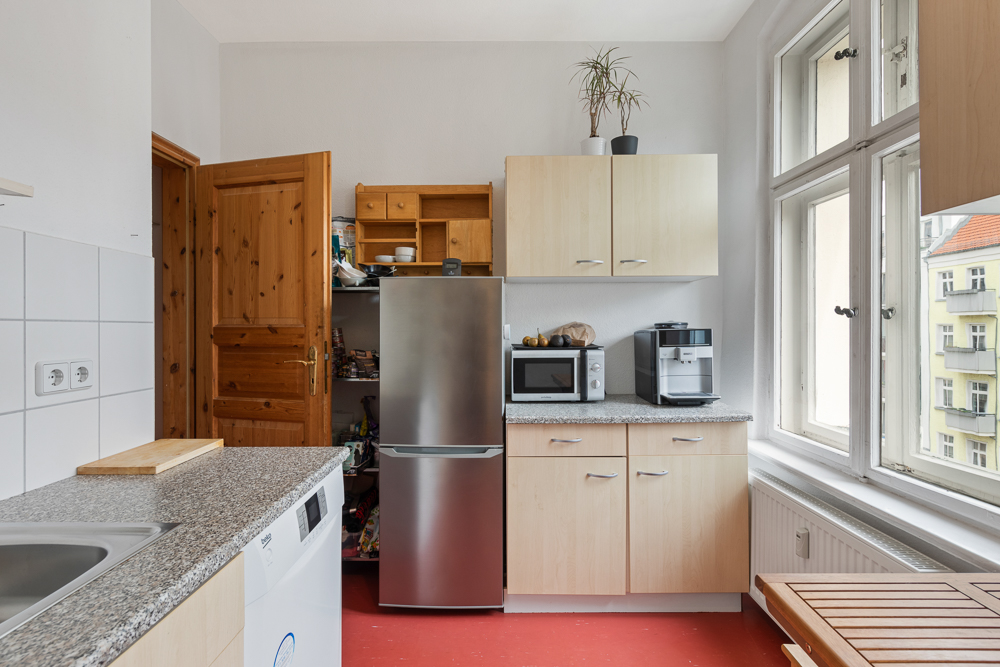 Helle Küche mit Kühlschrank, Oberschränken, Kaffeemaschine und großem Fenster.