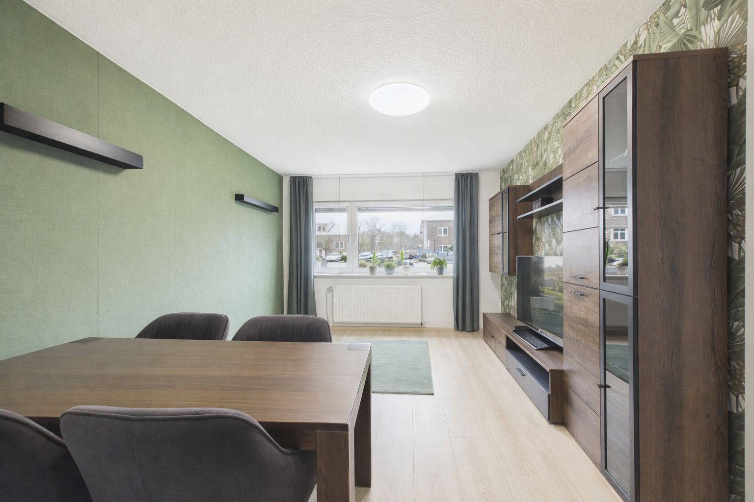 Wohnzimmer mit Esstisch, Stühlen, TV-Schrankwand, grün gestrichener Wand und großem Fenster.
