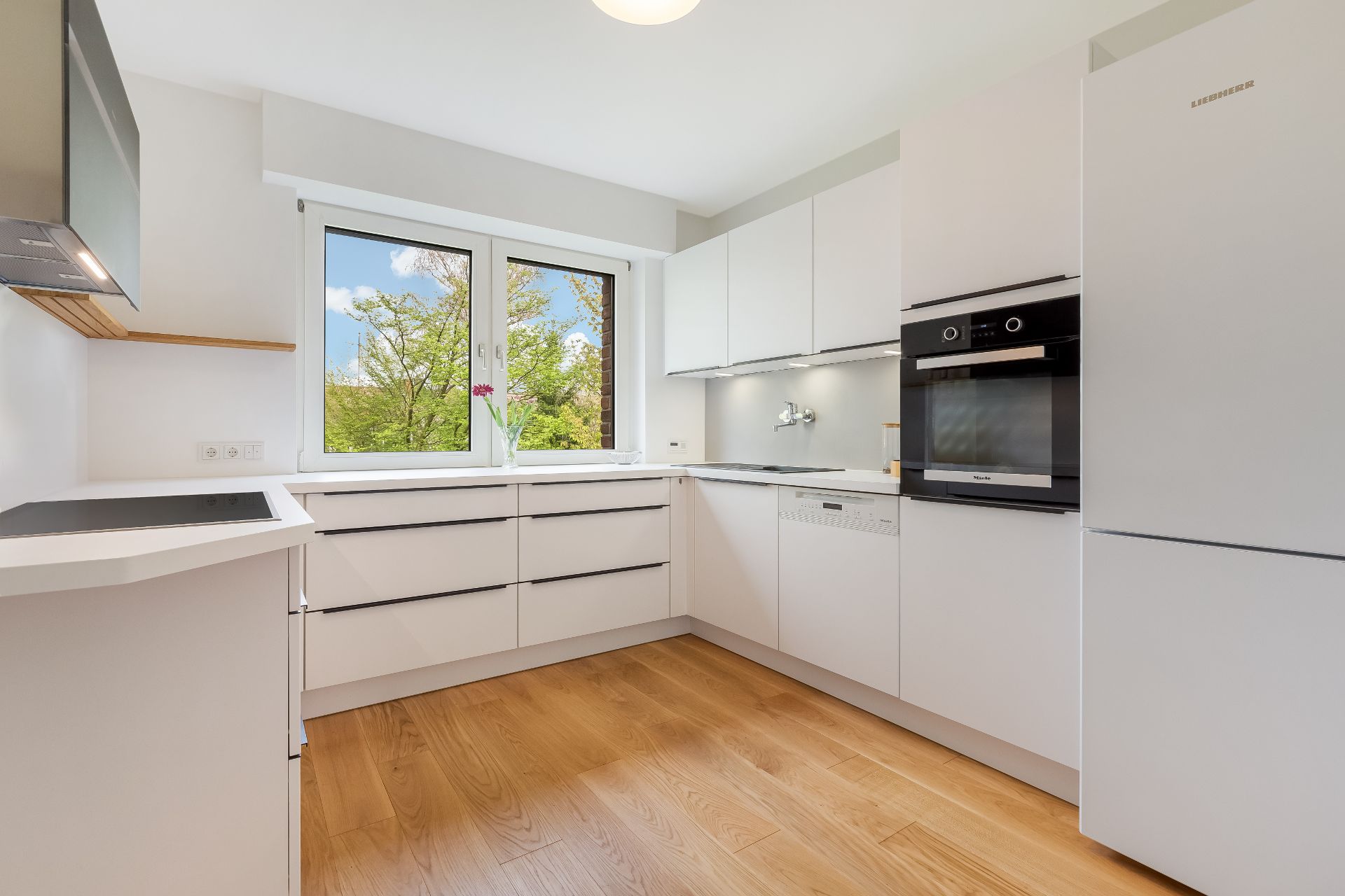 Moderne weiße Einbauküche mit Holzfußboden, großem Fenster und Geräten.