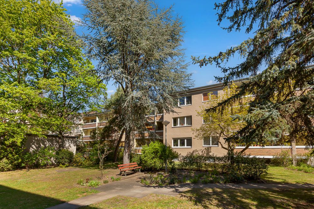Mehrfamilienhaus mit Garten, Bäumen und einer Parkbank im Vordergrund bei sonnigem Wetter.