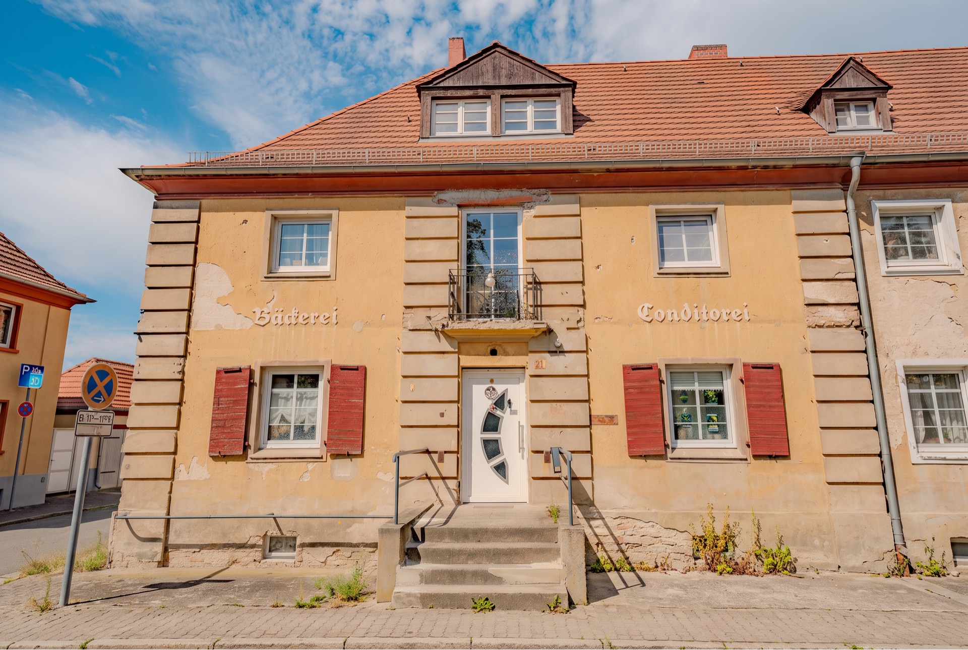 Altes Wohnhaus mit rotem Dach, cremefarbener Fassade und roten Fensterläden, zentrale Eingangstür.