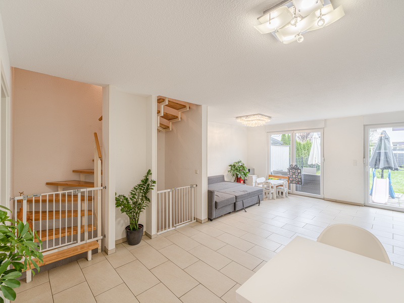 Helles Wohnzimmer mit Sofa, Kindertisch, Pflanzen, Treppe und Zugang zur Terrasse mit Garten.