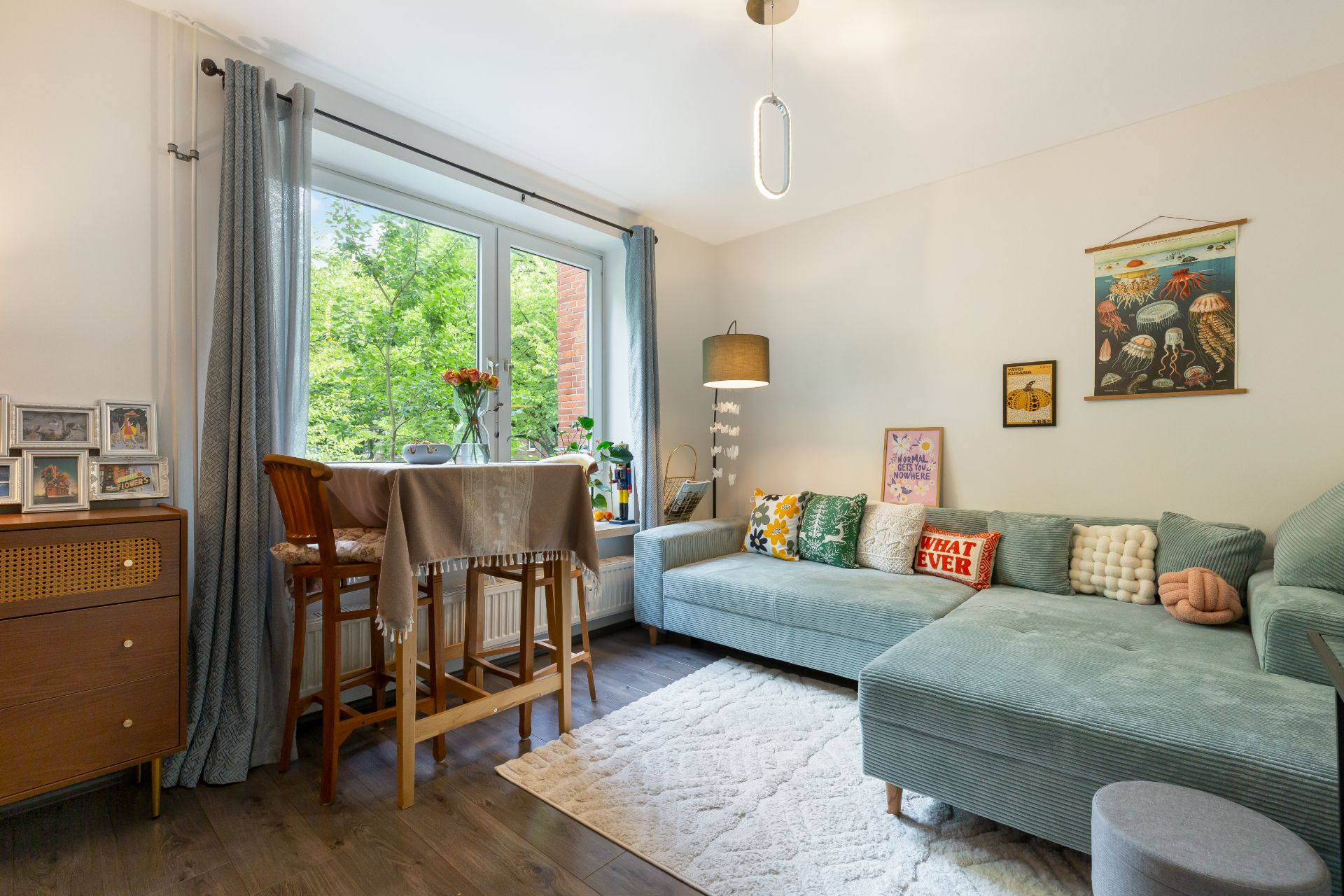 Helles Wohnzimmer mit Ecksofa, Teppich, Stehlampe, Esstisch und Bildern an der Wand.
