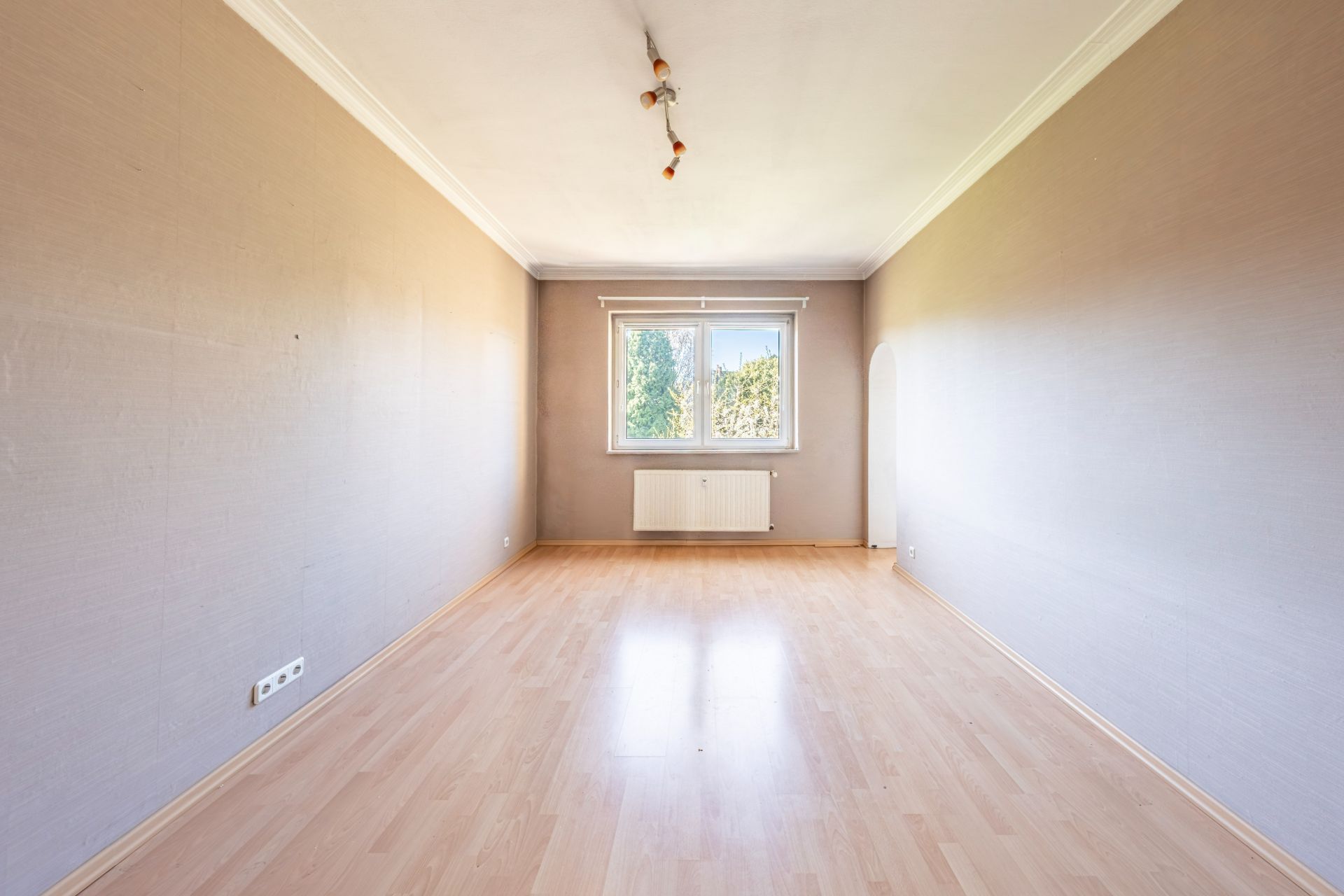 Leerer Raum mit hellem Laminatboden, Fenster und Heizkörper an der Stirnwand.