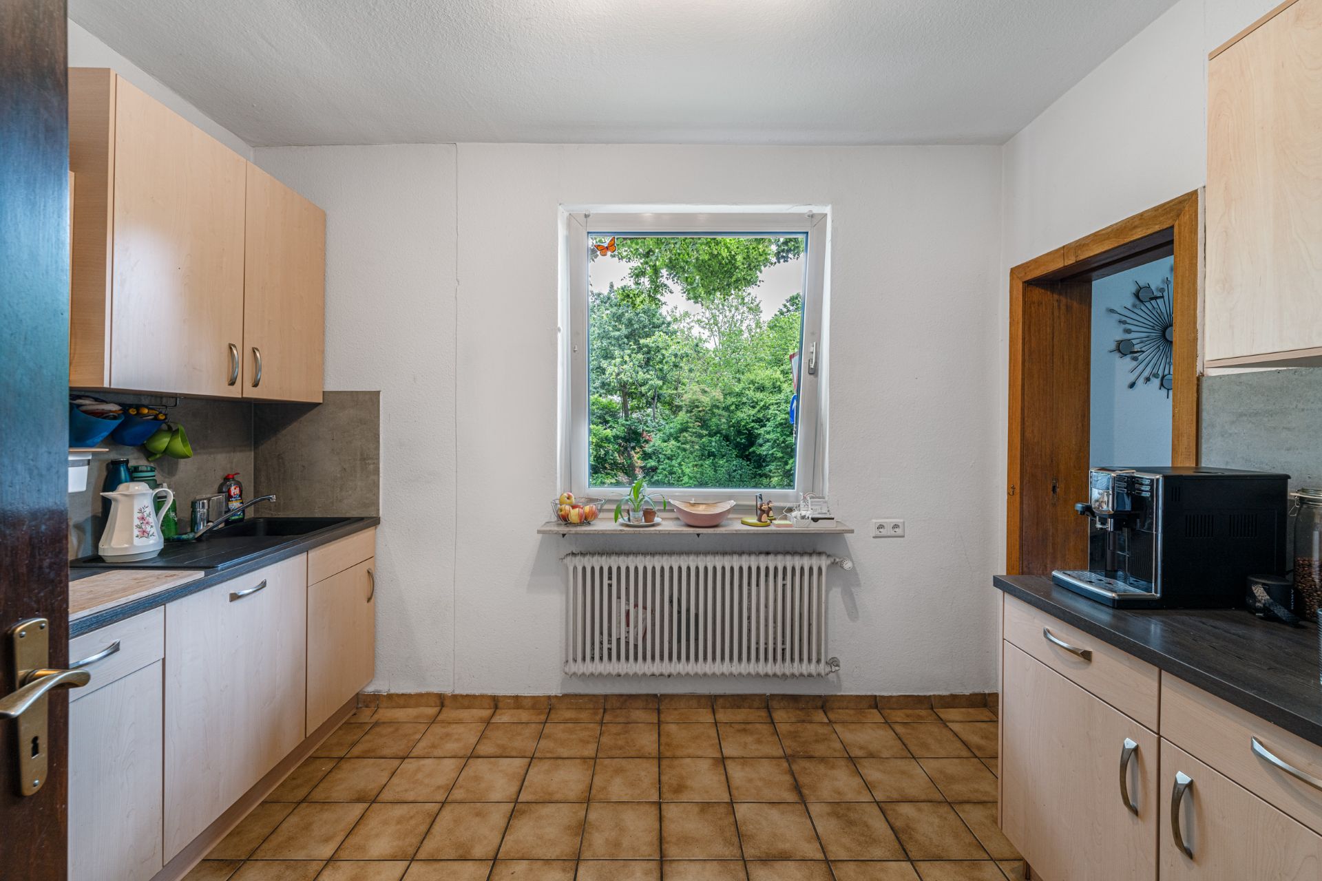 Helle Küche mit Holzmöbeln, Fenster mit Blick ins Grüne und Fliesenboden.