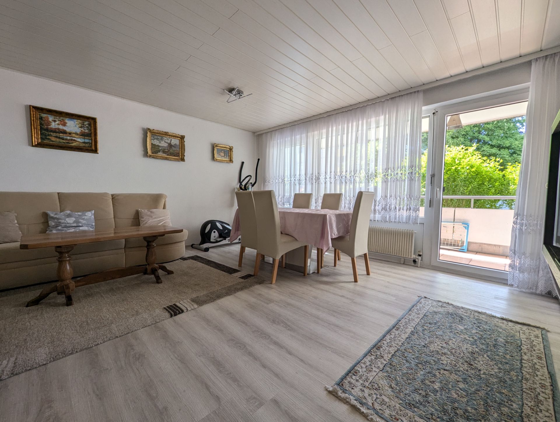 Wohnzimmer mit Sofa, Esstisch, vier Stühlen, Teppichen und Zugang zu einem Balkon mit Glasfront.