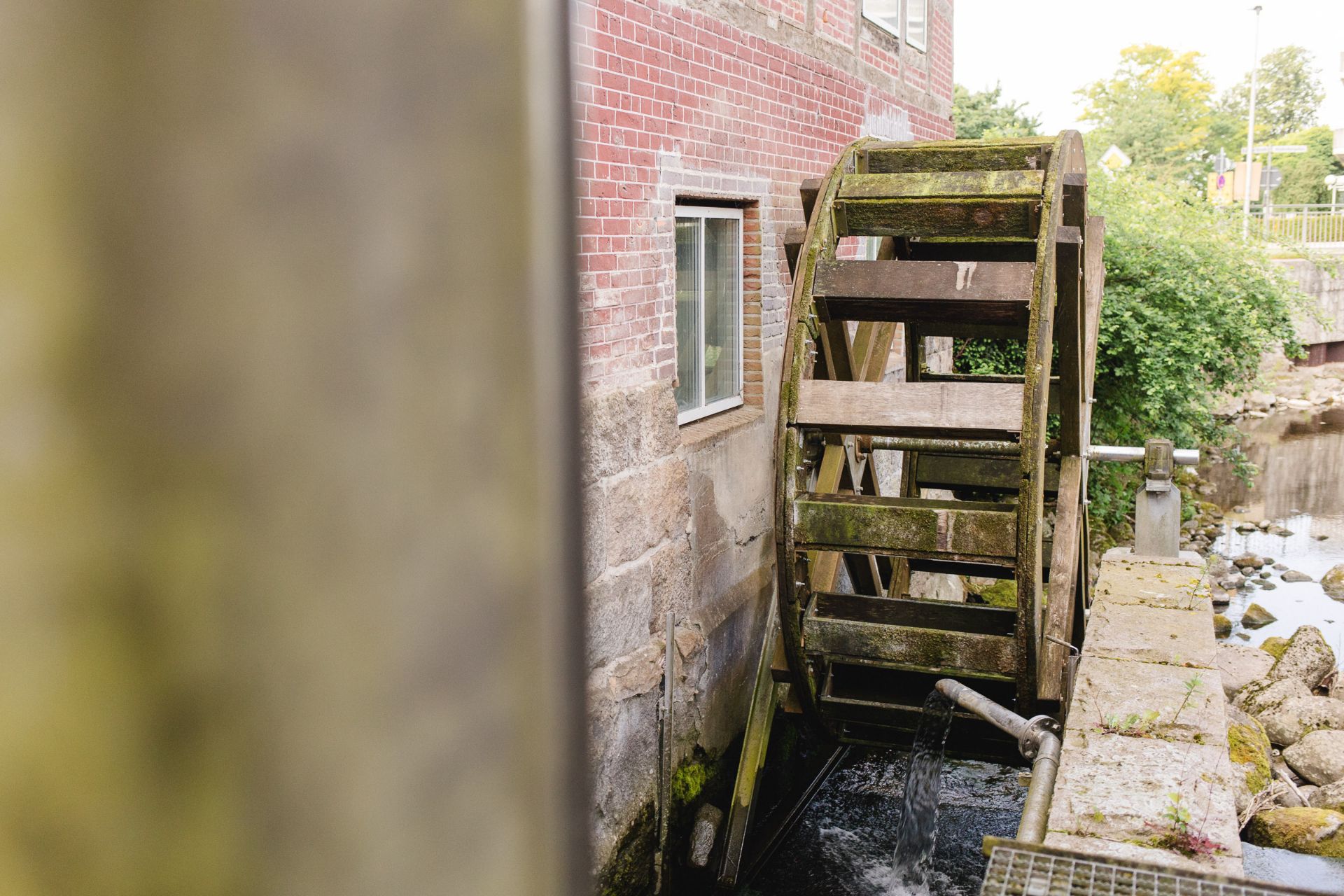 Holz-Wasserrad an einer Backsteinmauer mit Fenster, Wasser fließt darunter.