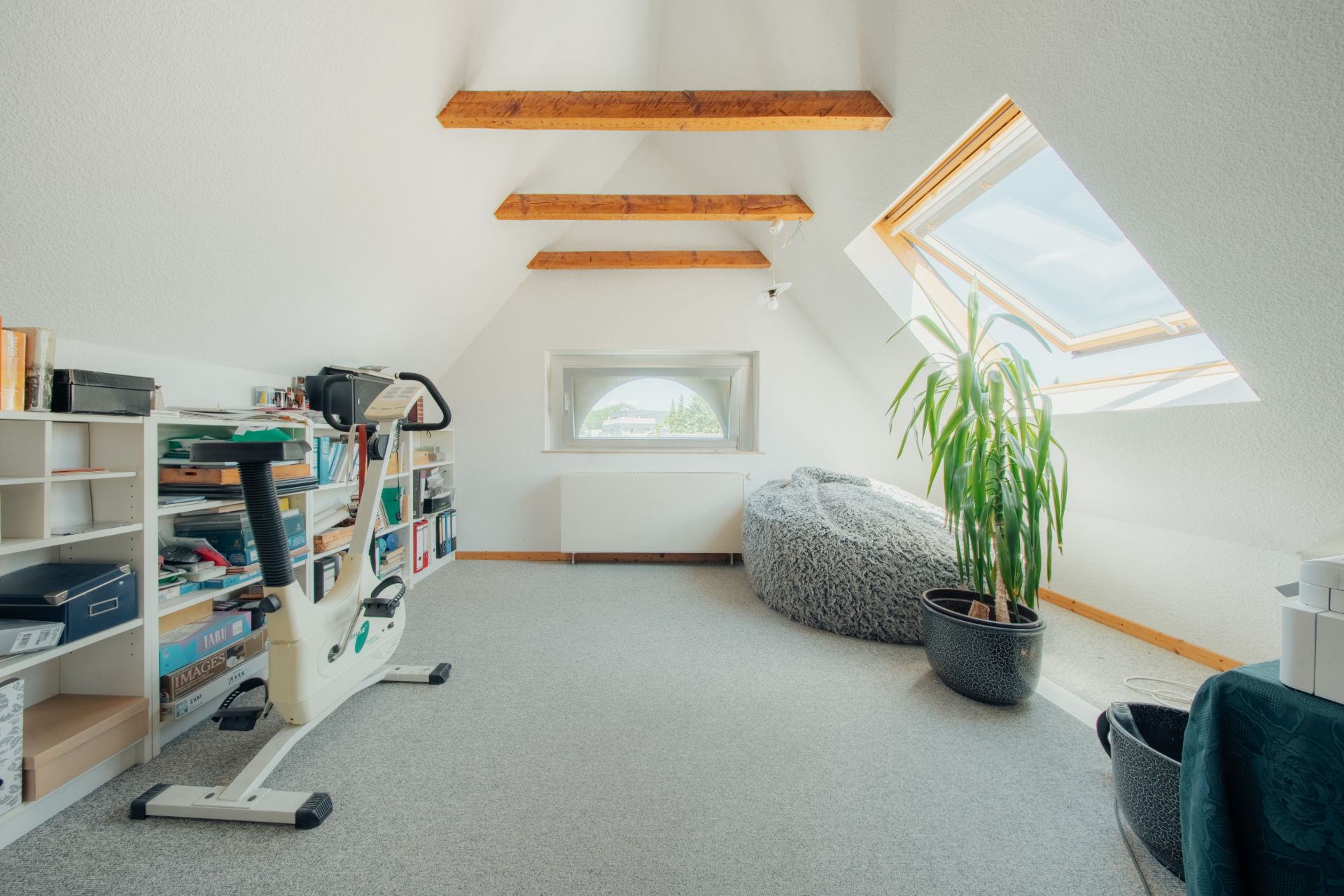 Heller Dachbodenraum mit Bücherregal, Heimtrainer, Pflanze und Sitzsack unter Dachfenster.