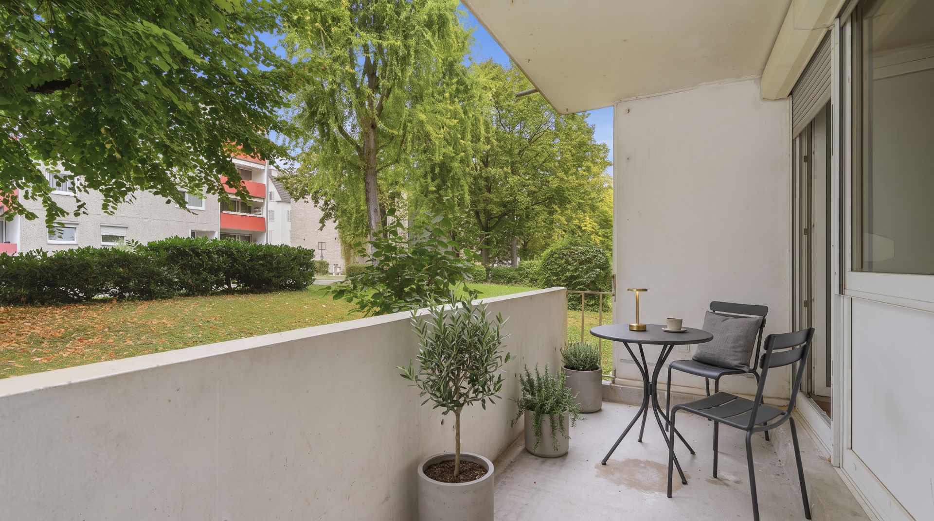 Balkon mit zwei Stühlen, Tisch und Topfpflanzen, Blick auf grüne Bäume und Wiese.