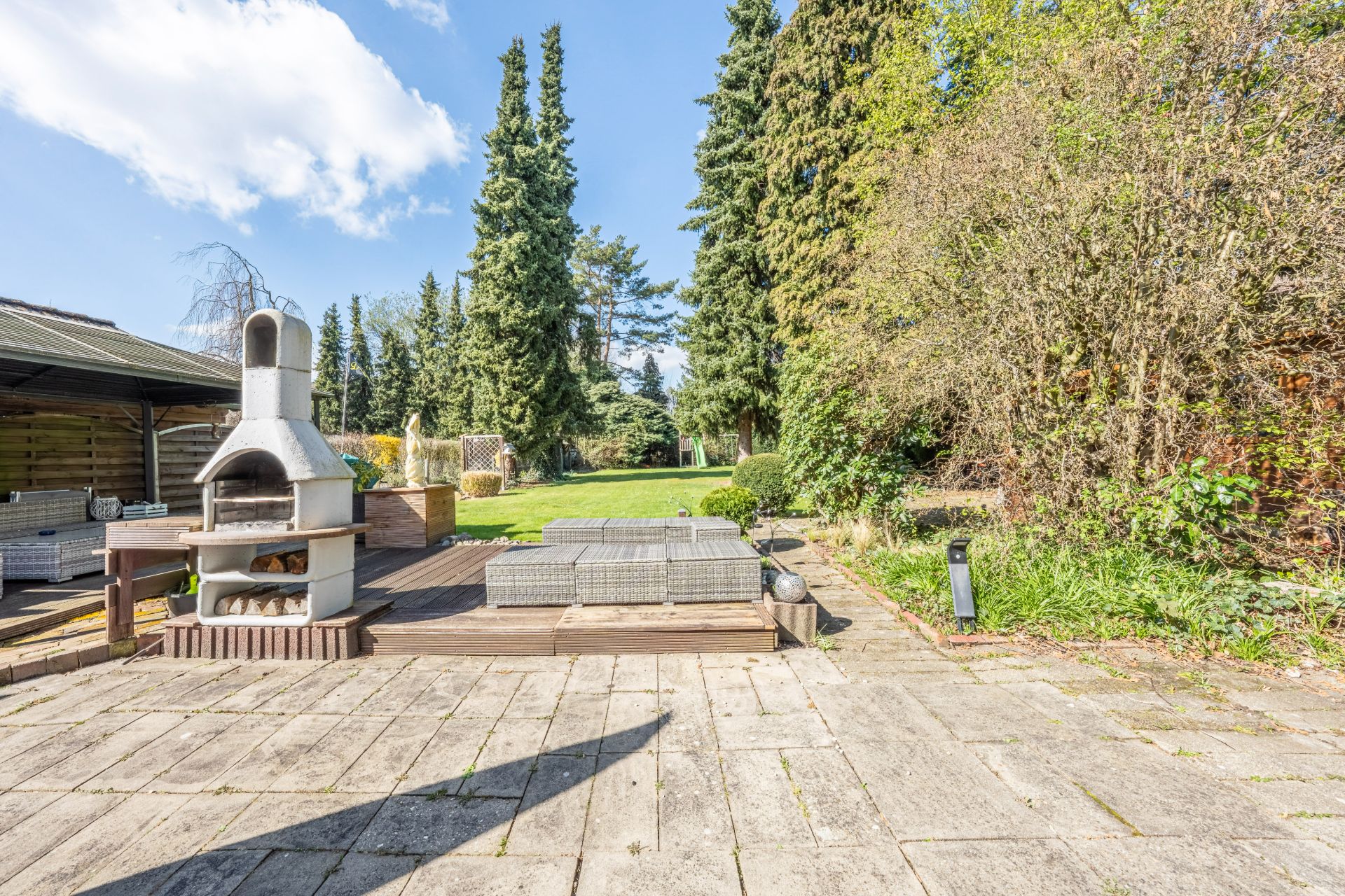 Gepflasterte Terrasse mit Steinofen und Gartenmöbeln, dahinter Rasenfläche und hohe Bäume.