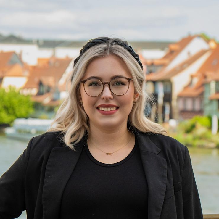 Junge Frau mit Brille und schwarzer Kleidung steht draußen vor Häusern mit roten Dächern.