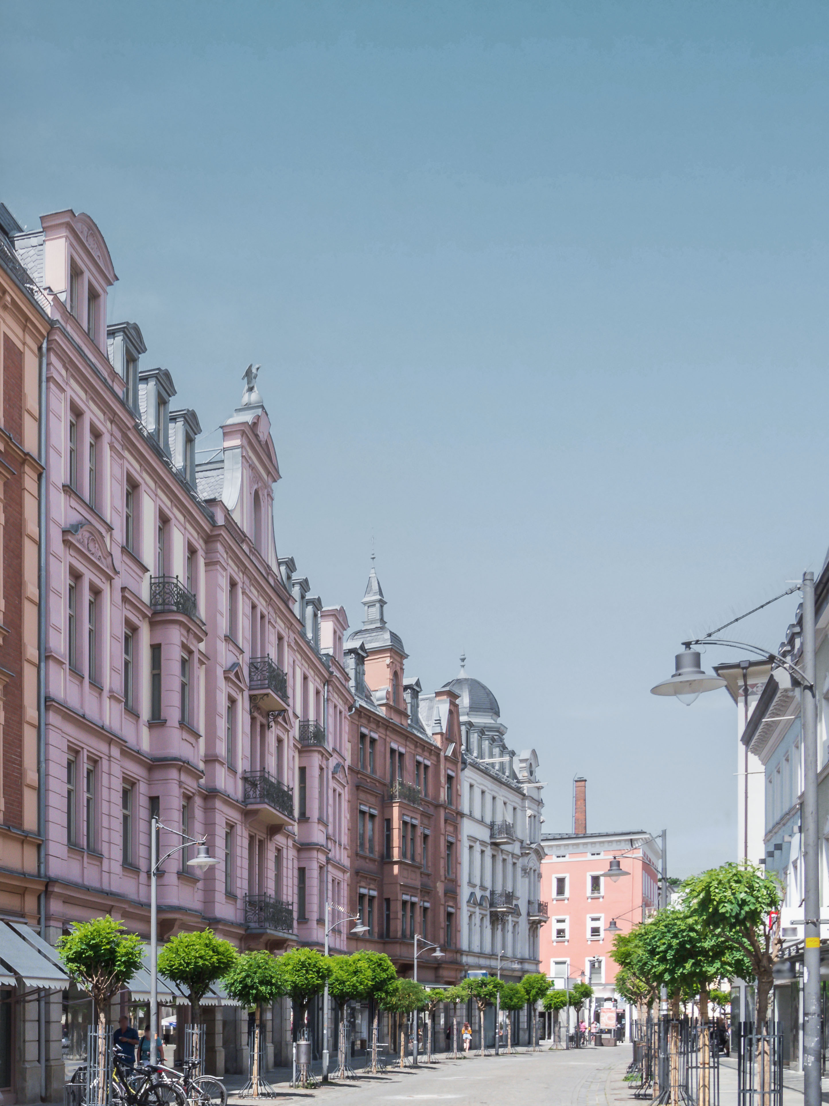 Straße mit mehrstöckigen Altbauten und kleinen Bäumen am Gehweg.
