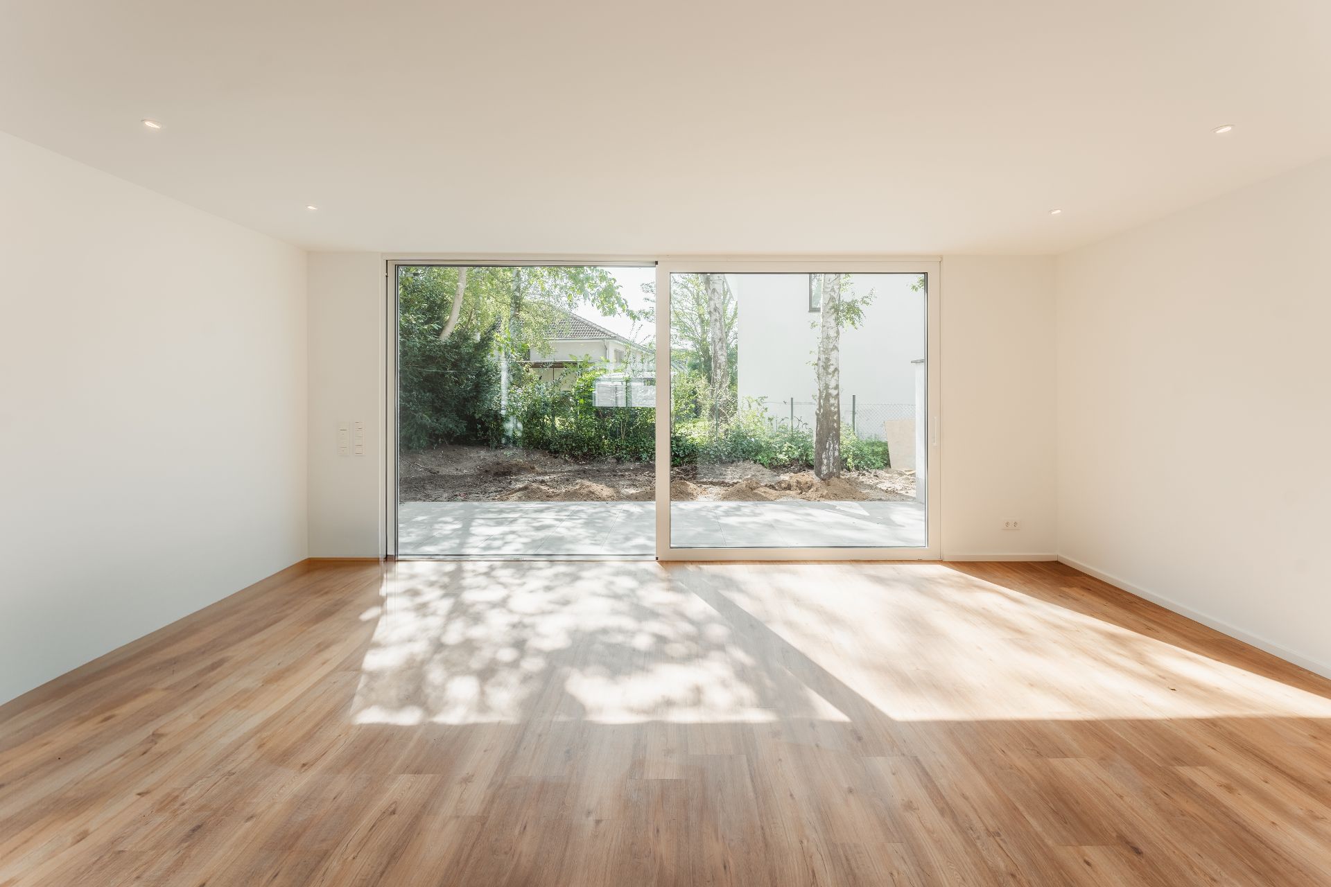 Heller leerer Raum mit Holzfußboden und großer Glas-Schiebetür zum Garten.