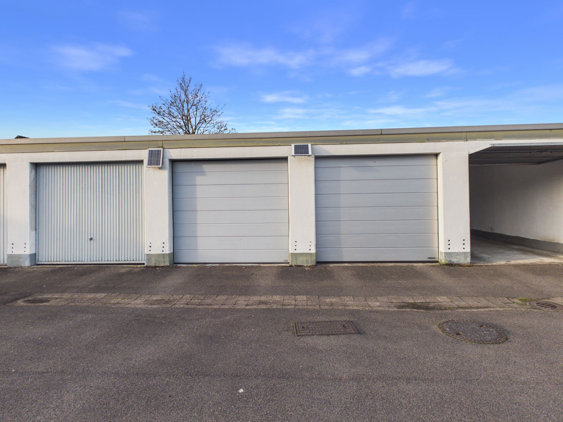 Drei geschlossene Garagentore und eine offene Garage nebeneinander auf einem Parkplatz.