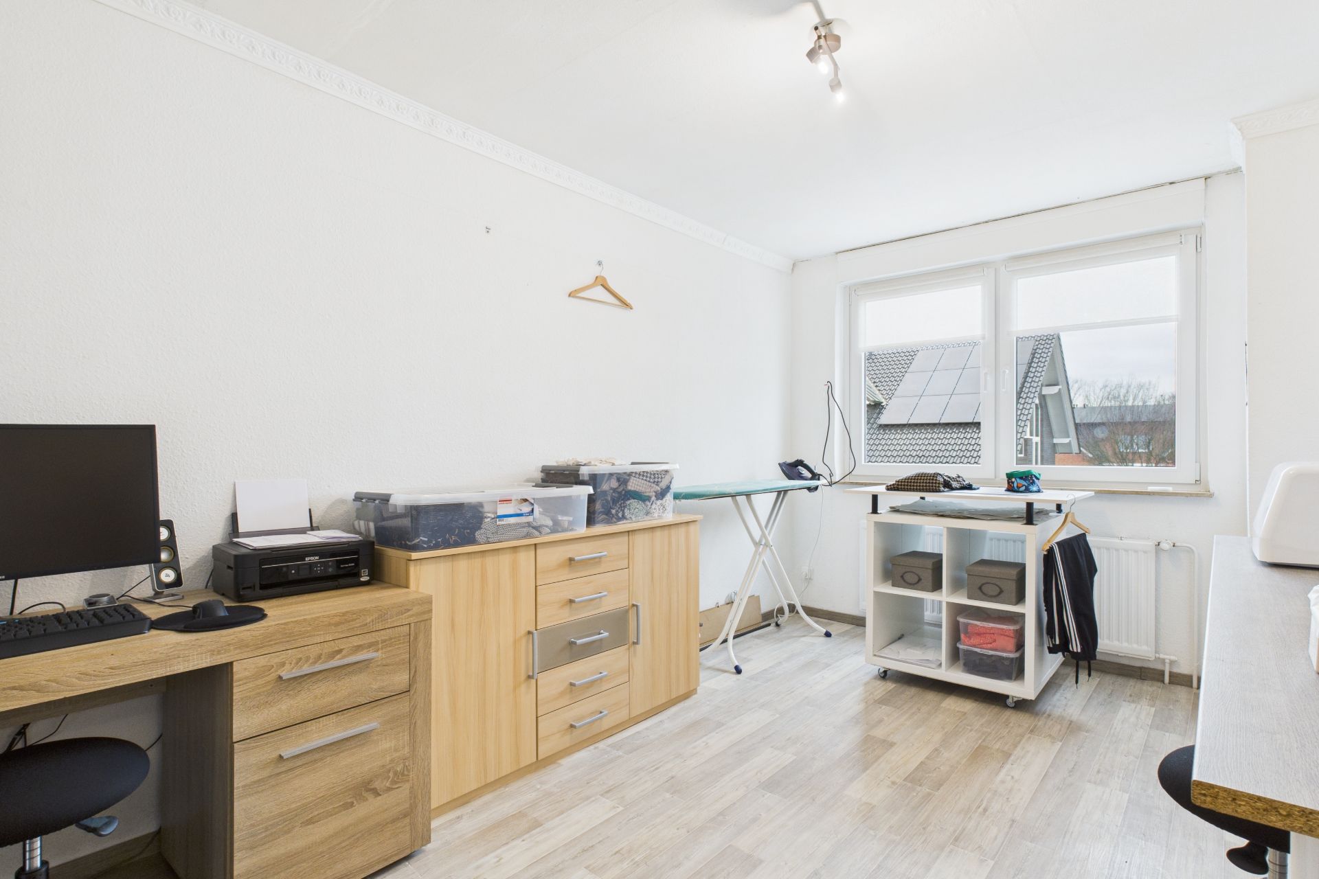 Helles Arbeitszimmer mit Schreibtisch, Computer, Kommode und großem Fenster.