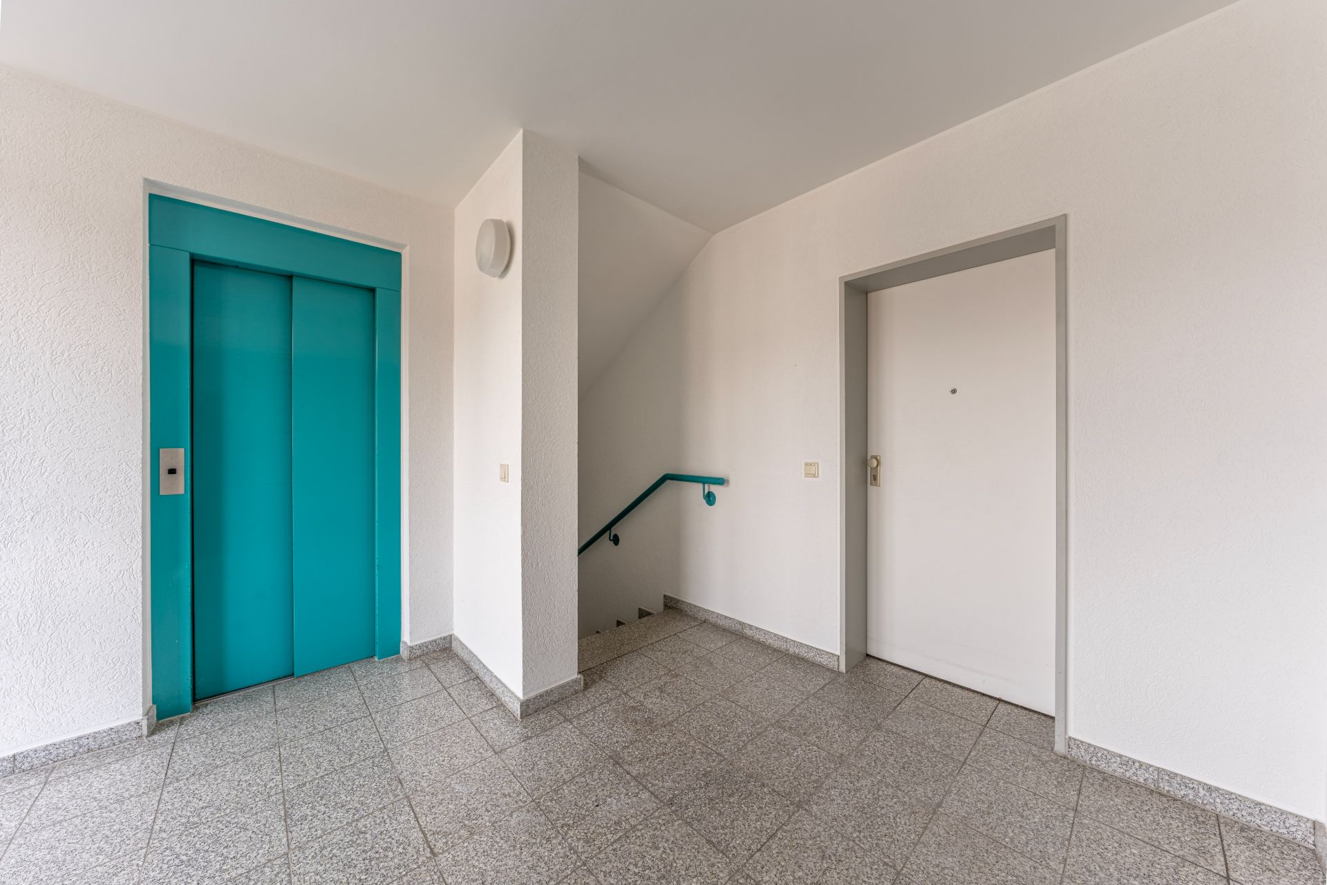 Hausflur mit türkisfarbenem Aufzug, Treppenhaus und weißer Wohnungstür.