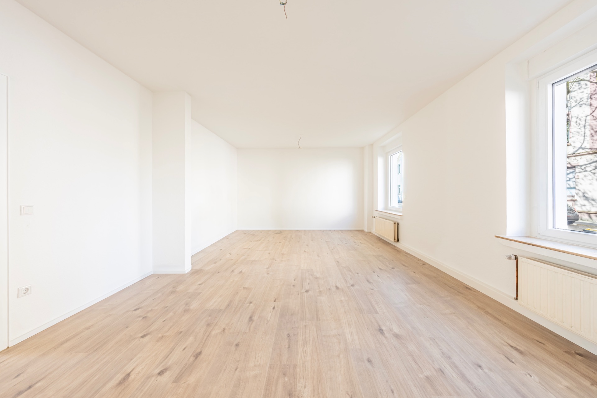 Helles, leeres Zimmer mit Holzfußboden und großen Fenstern an der rechten Wand.