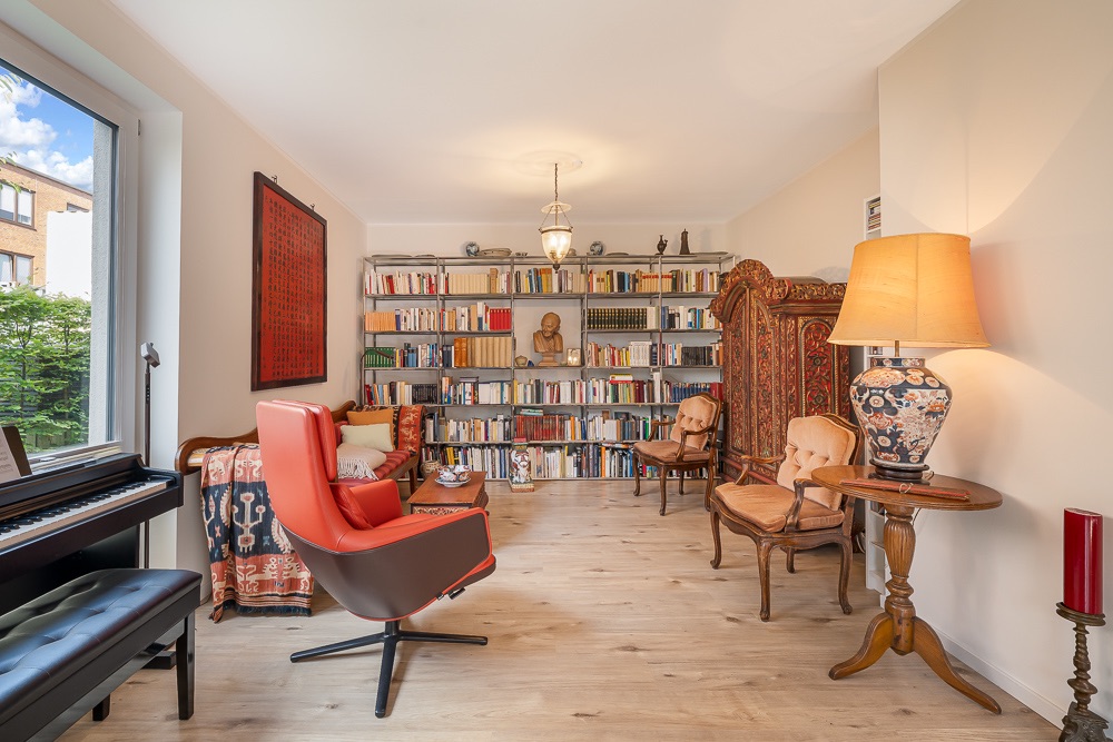Wohnzimmer mit Bücherregal, roten Sesseln, Stehlampe, Klavier und großem Fenster.