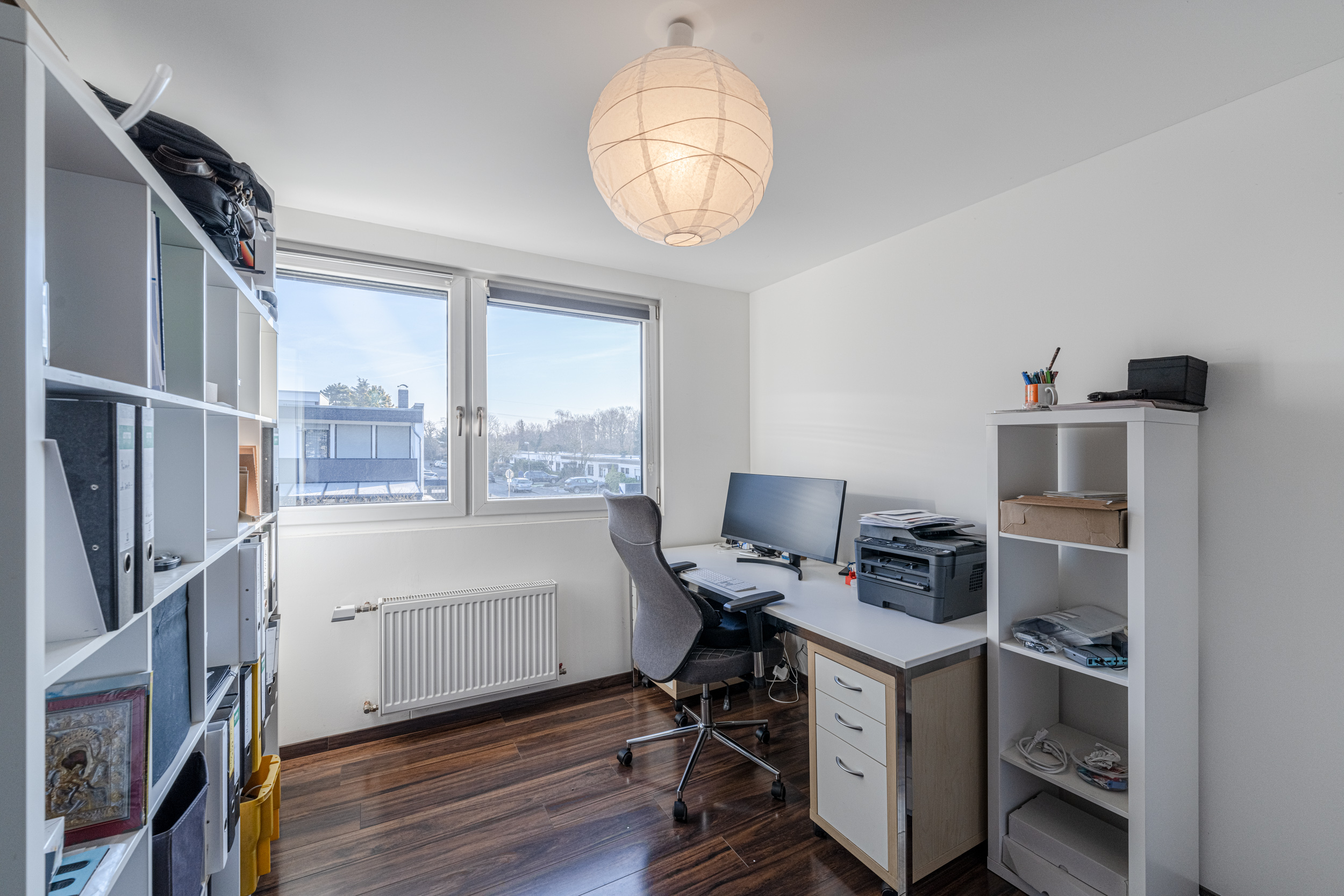 Helles Arbeitszimmer mit Schreibtisch, Monitor, Regal, Stuhl und großem Fenster.